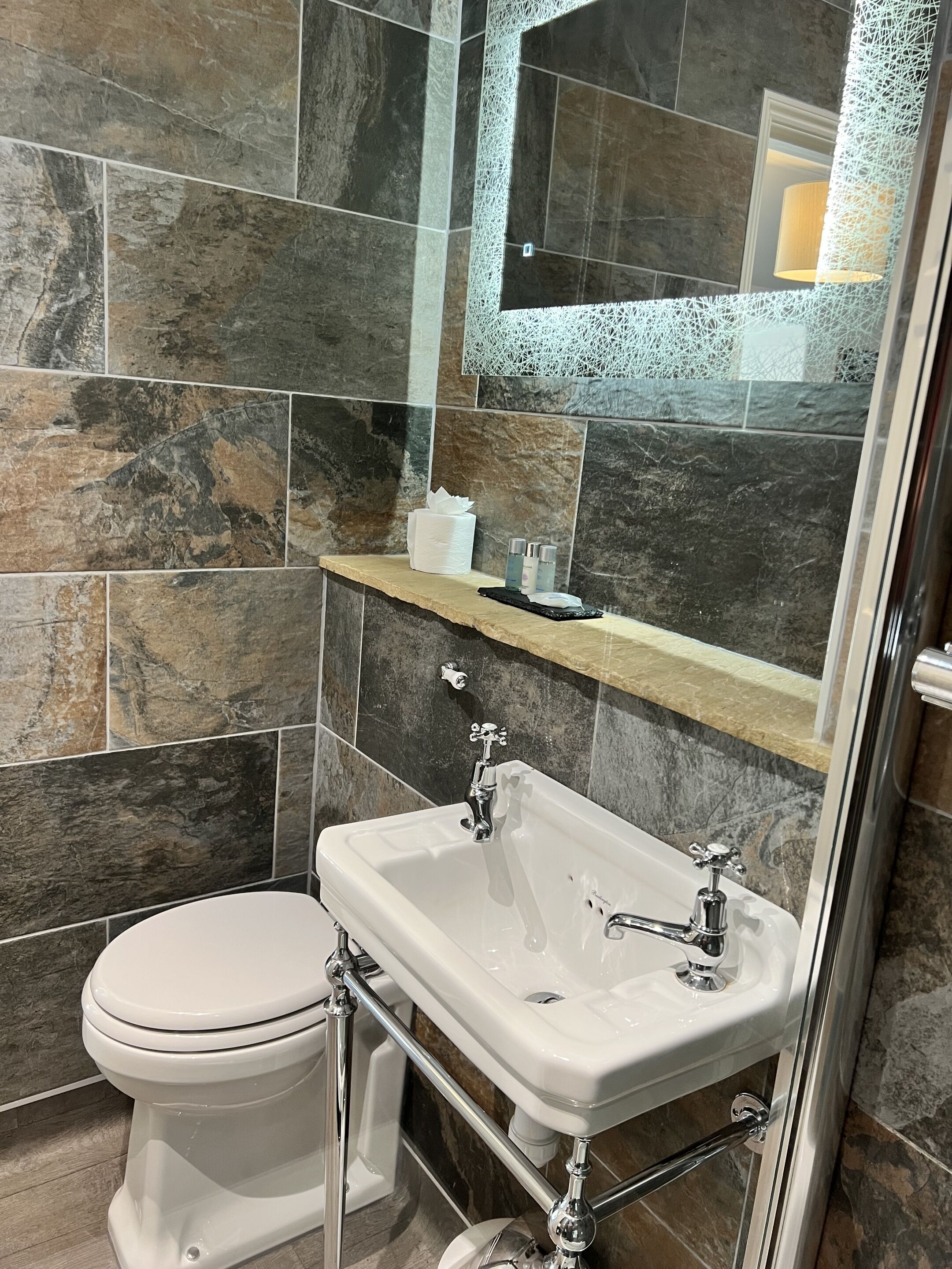 A bathroom with a white toilet, a white sink with chrome fixtures, and a mirror above the sink. The walls are tiled with dark stone-like tiles.