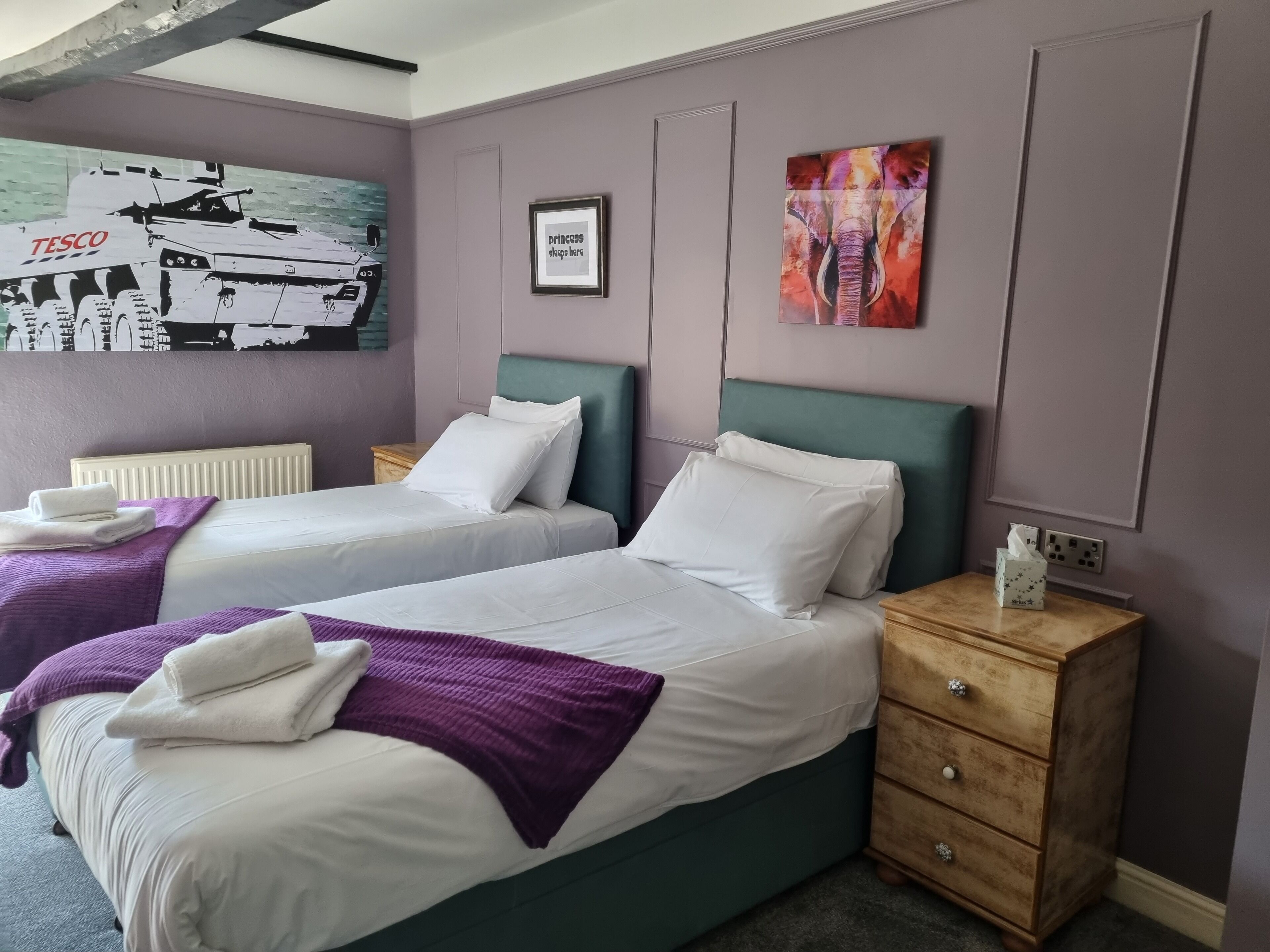 A hotel room with two beds, purple bedding, a green headboard, a wooden nightstand with a tissue box, a radiator, and artwork on the walls.