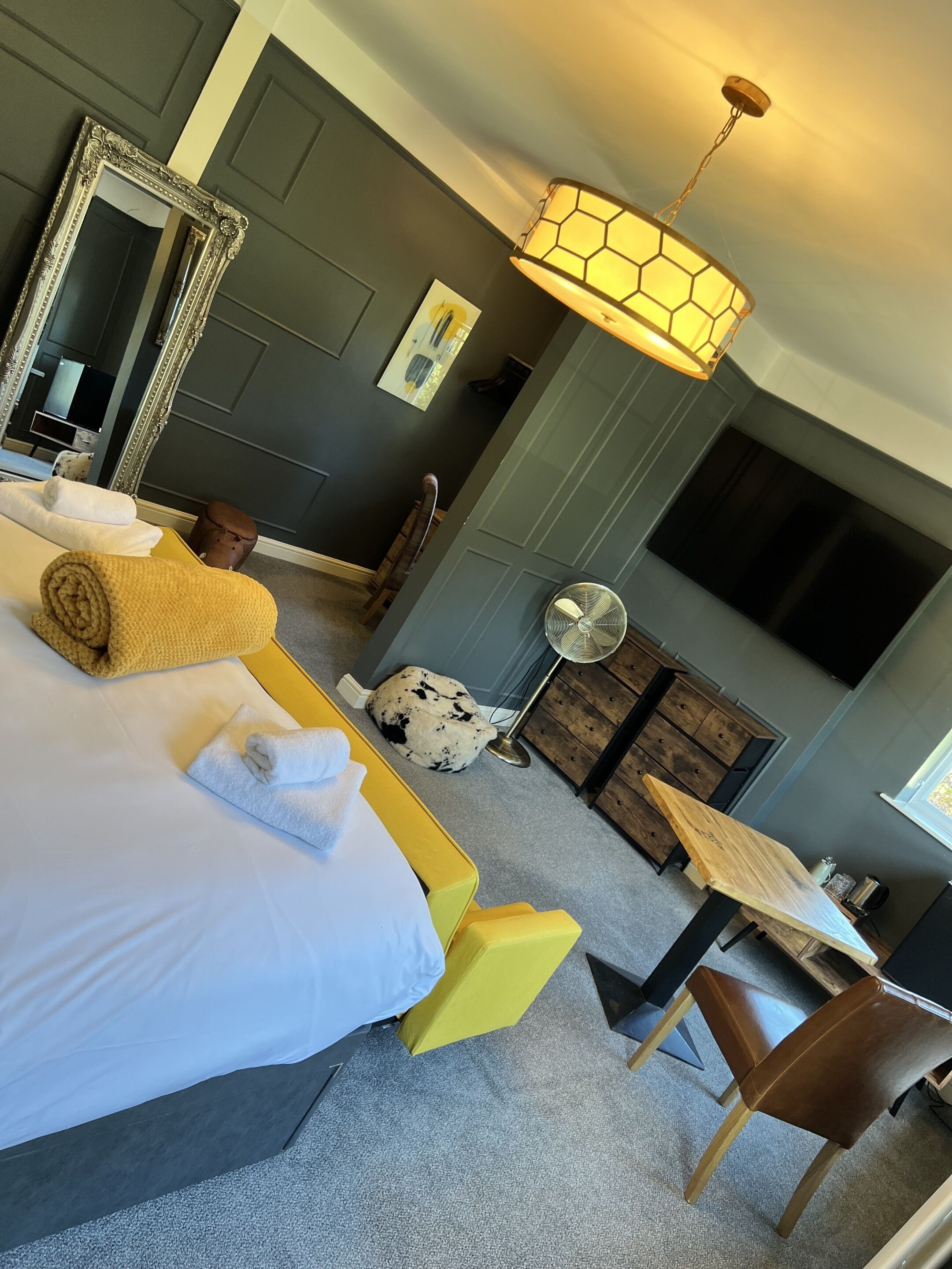 A hotel room with a bed, a desk, a chair, a mirror, a fan, and a dresser.