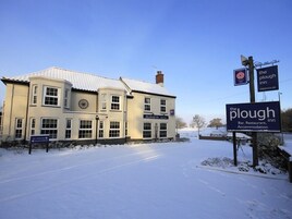 A two-story building with a snow-covered roof and a signboard for 'The Plough Inn' indicating it is a bar, restaurant, and accommodation.