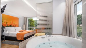 Un dormitorio moderno con una bañera grande, una cama con ropa blanca y vistas a zonas verdes por la ventana.