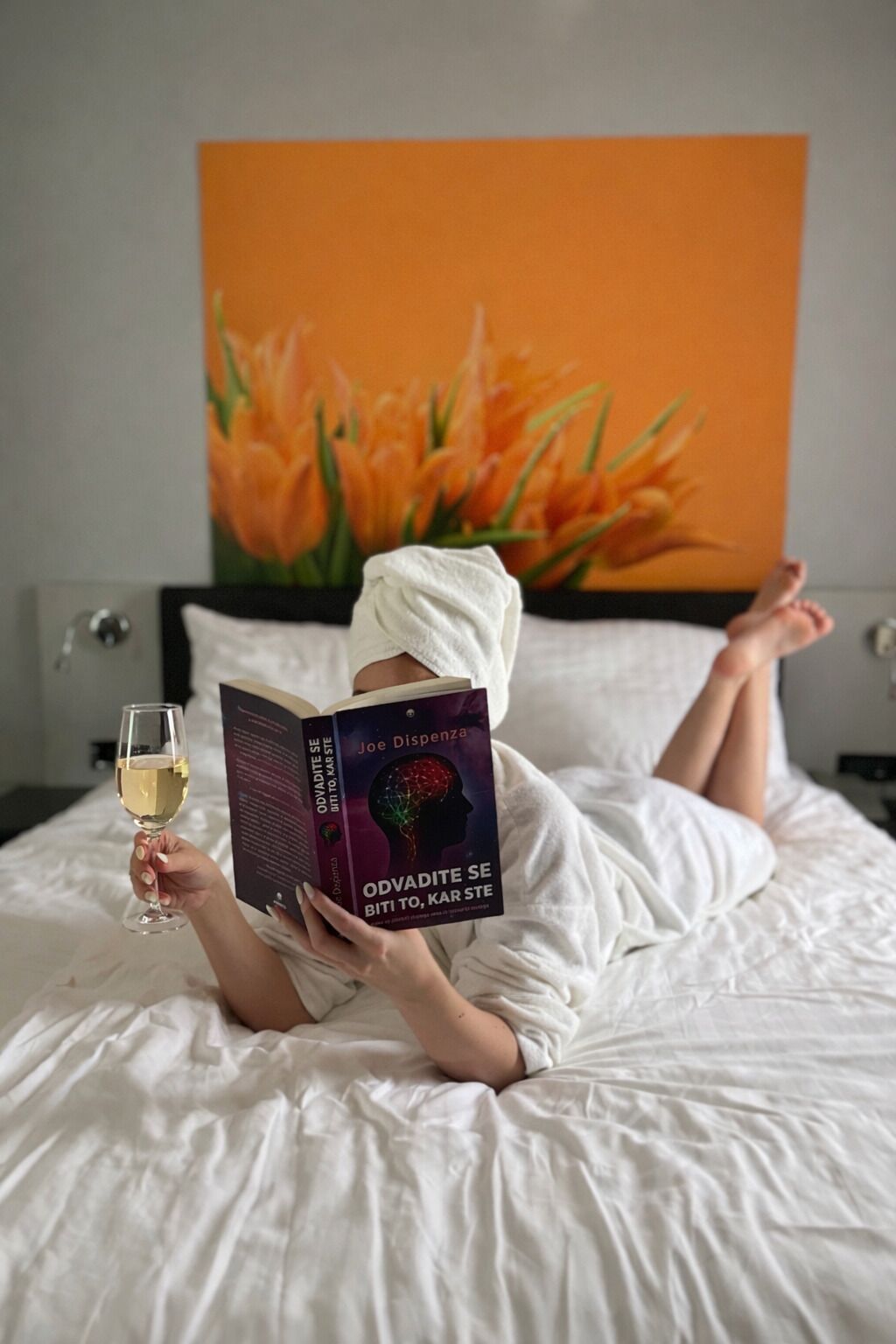 A person lying on a bed reading a book with a glass of wine, with a large flower painting in the background.