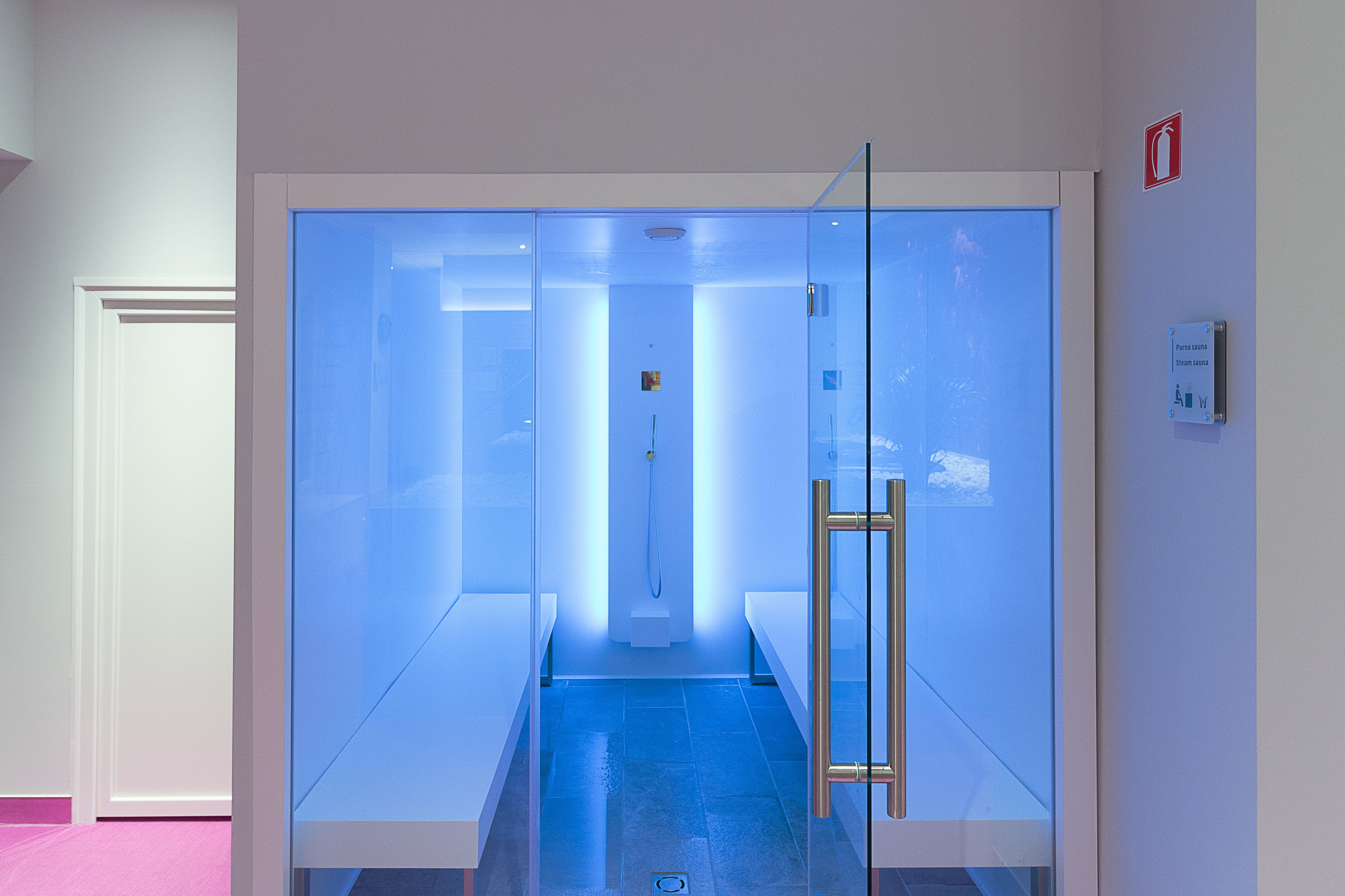 A modern, glass-walled sauna with a blue-lit interior, a gold handle, and a visible showerhead.