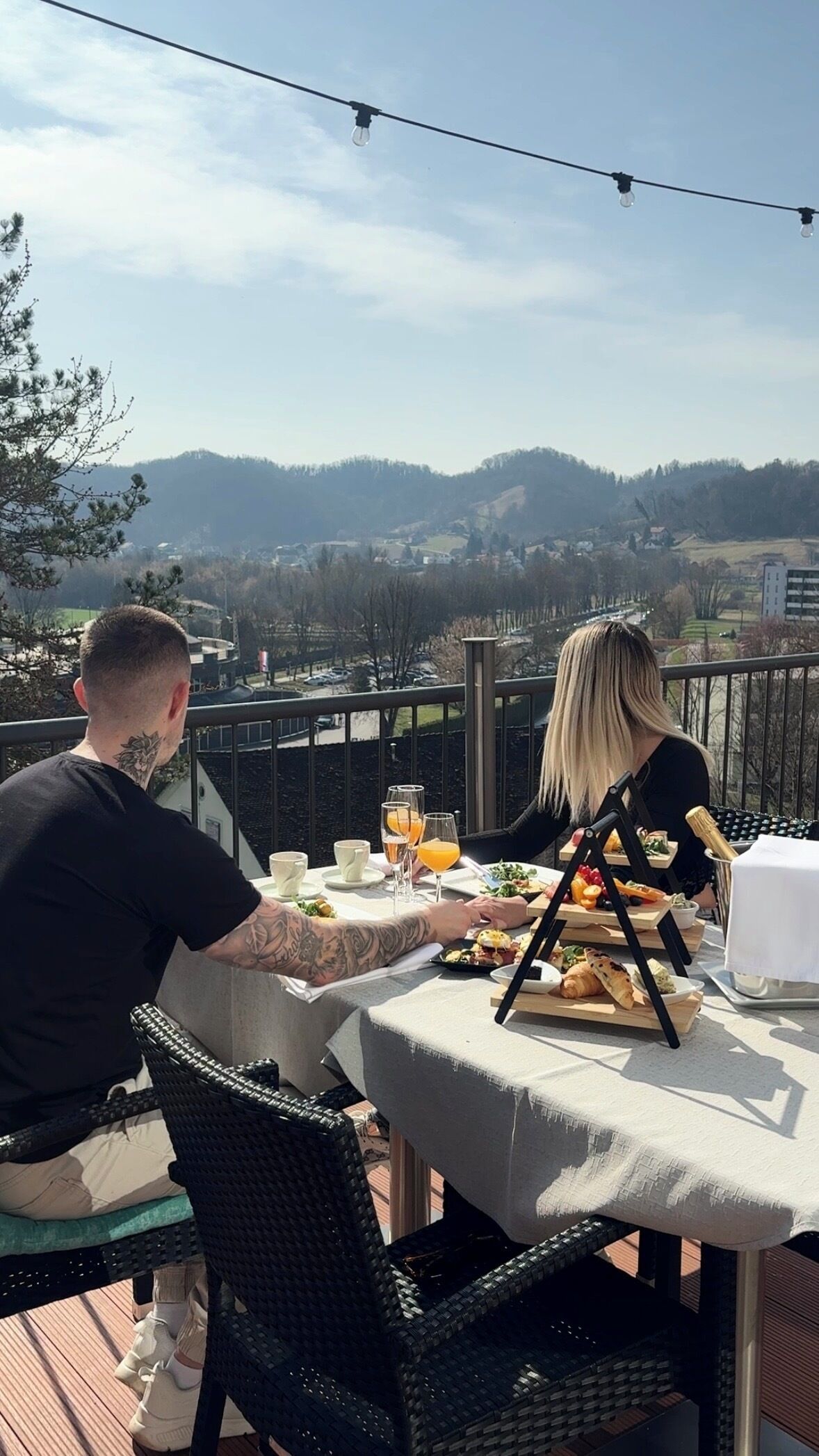 Un couple est assis à une table pour deux, en plein air, et profite d’une vue panoramique sur des collines et une ville lointaine.