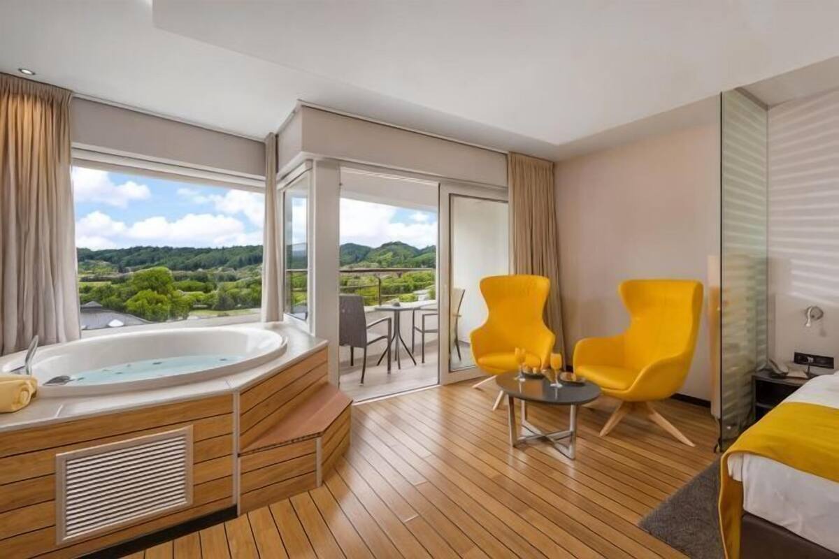 Une chambre d’hôtel avec un plancher en bois, une grande fenêtre, une baignoire, deux chaises jaunes et un lit recouvert d’une courtepointe blanche et jaune.