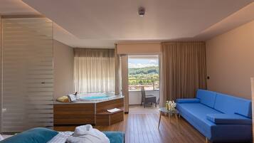 A hotel room with a bed, a blue sofa, a wooden desk with a laptop, a window with curtains, and a balcony with a view of greenery.