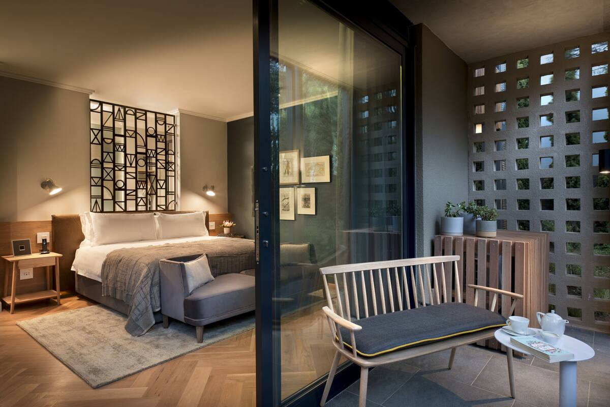 A modern bedroom with a bed, bedside tables, a bench, a small table with a teapot, and a glass partition with artwork.