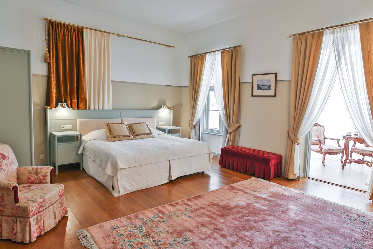 A bedroom with a bed, nightstands, a chair, a bench, and a view of the outside.