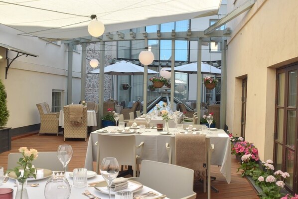 An outdoor dining area with tables set for two, surrounded by wicker chairs and covered by umbrellas.