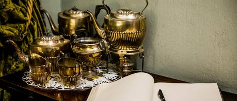 A table with a set of brass tea pots and cups, an open book, and a pen on a doily.