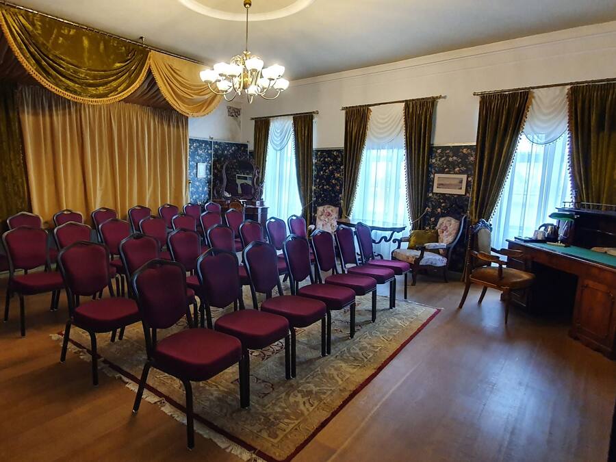 Um quarto com fileiras de cadeiras vermelhas, uma escrivaninha de madeira e um espelho à esquerda.