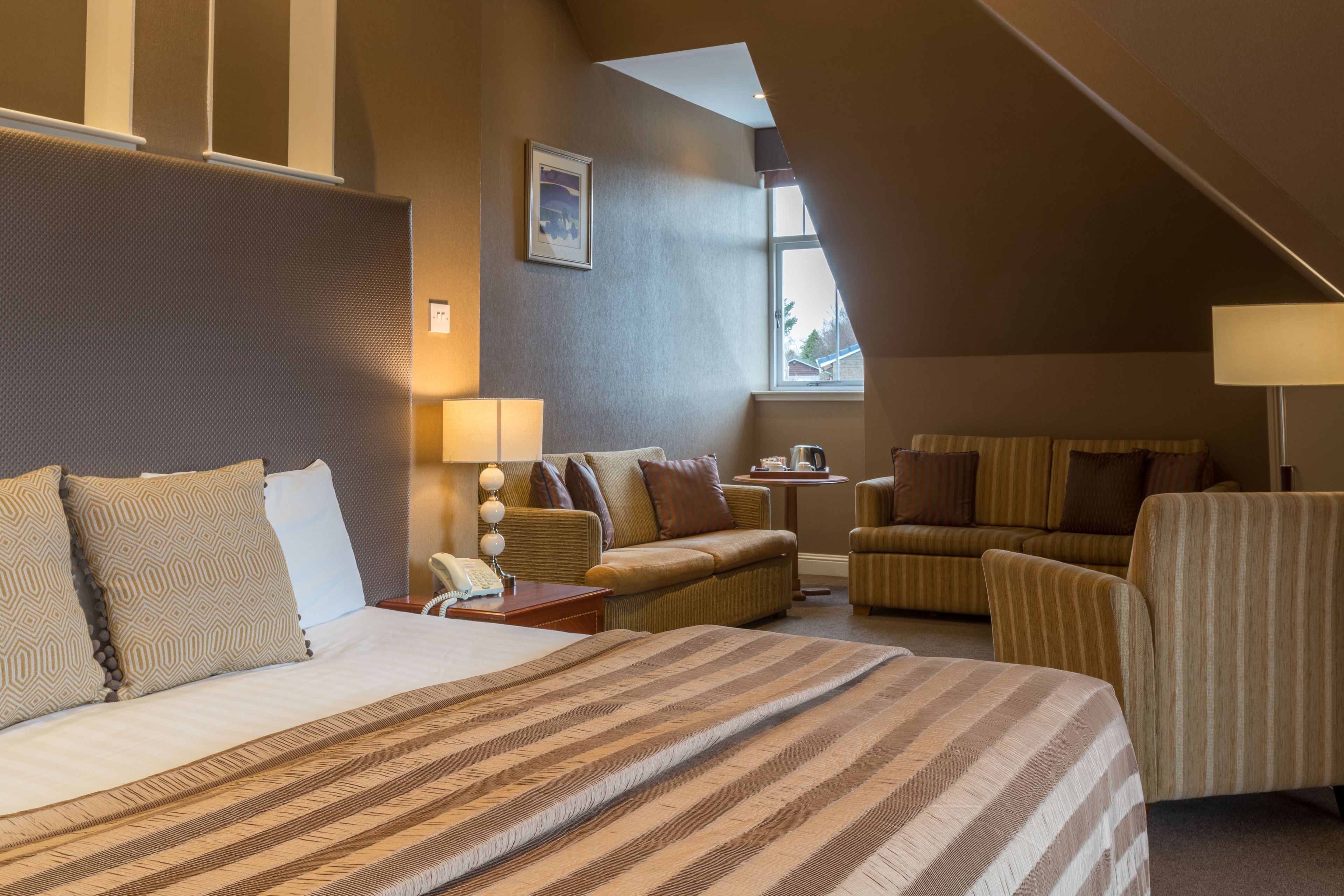 A hotel room with a large bed, two armchairs, a sofa, a small table with a lamp, and a window providing natural light.