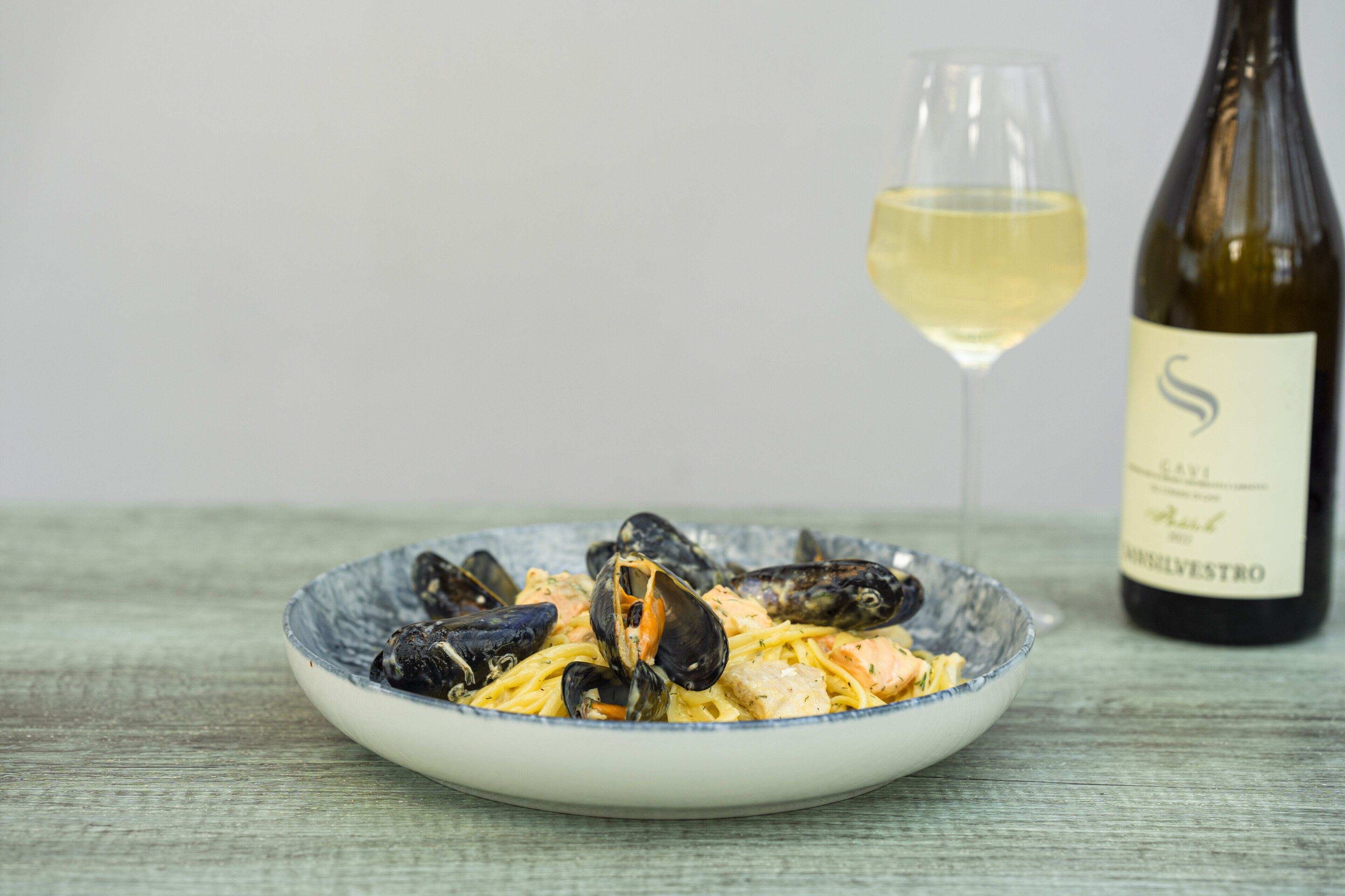 Un plato de mejillones con pasta, una copa de vino blanco y una botella de vino sobre una mesa de madera.