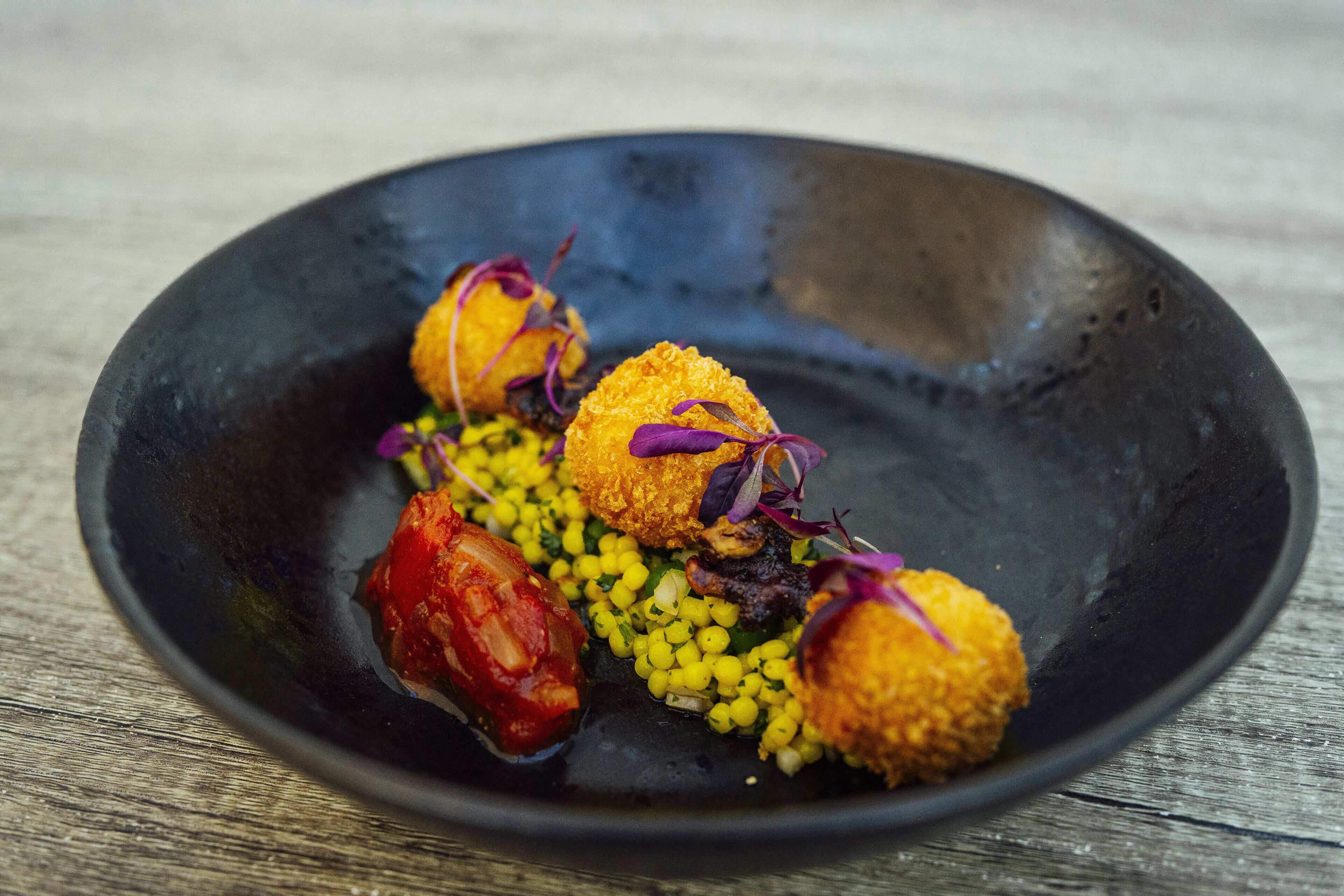 A black plate with three golden-brown fried items, some purple garnish, and yellow corn kernels.