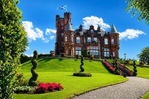 Un edificio grande e histórico con múltiples torres y una bandera, rodeado de jardines cuidados y un sendero de grava.