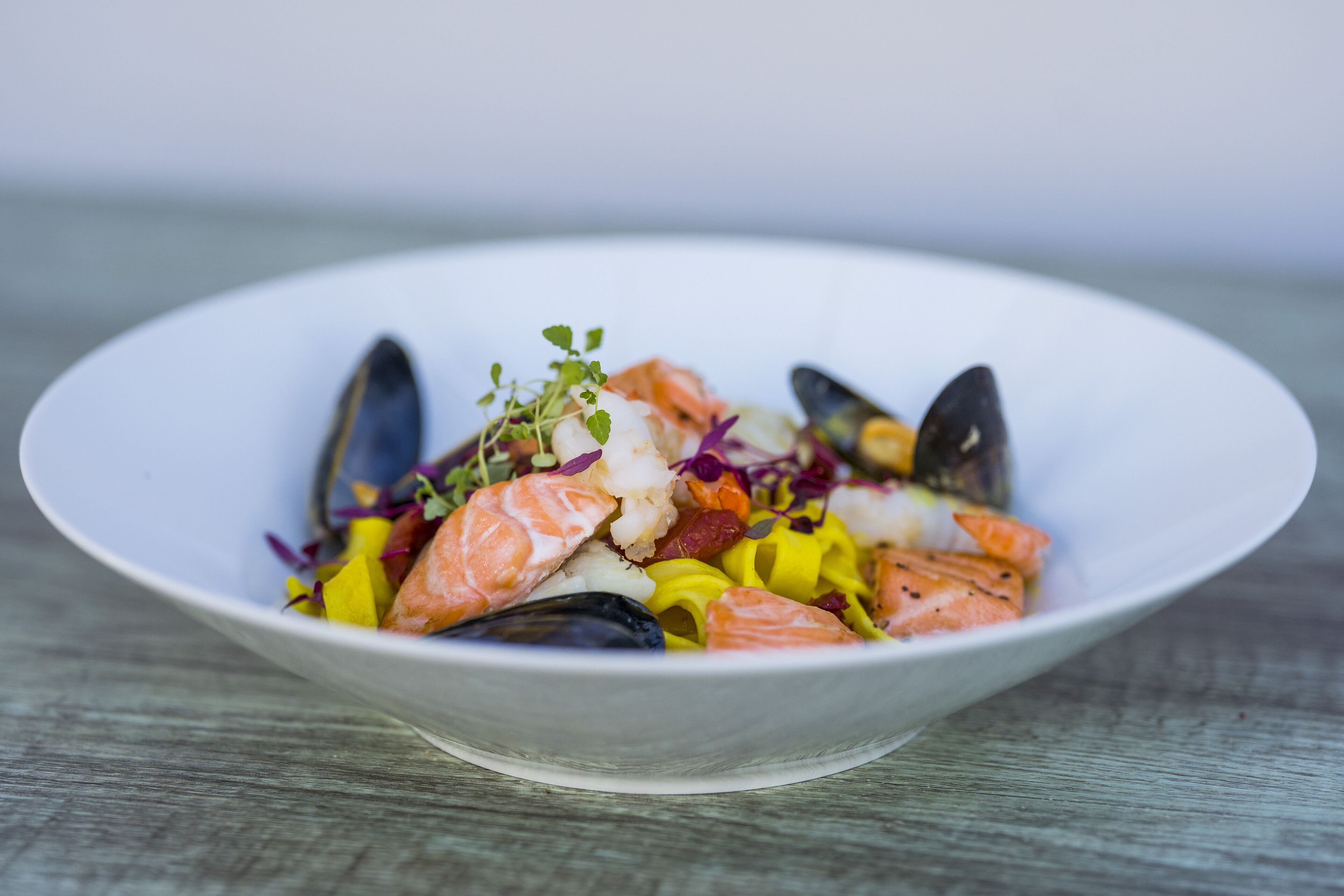 A white bowl containing a seafood dish with mussels, salmon, and vegetables.