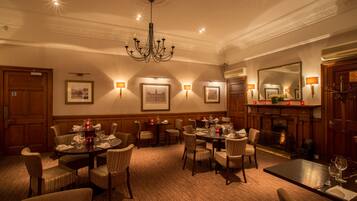 A well-lit dining area with multiple tables and chairs, a chandelier, framed pictures on the walls, and a fireplace.