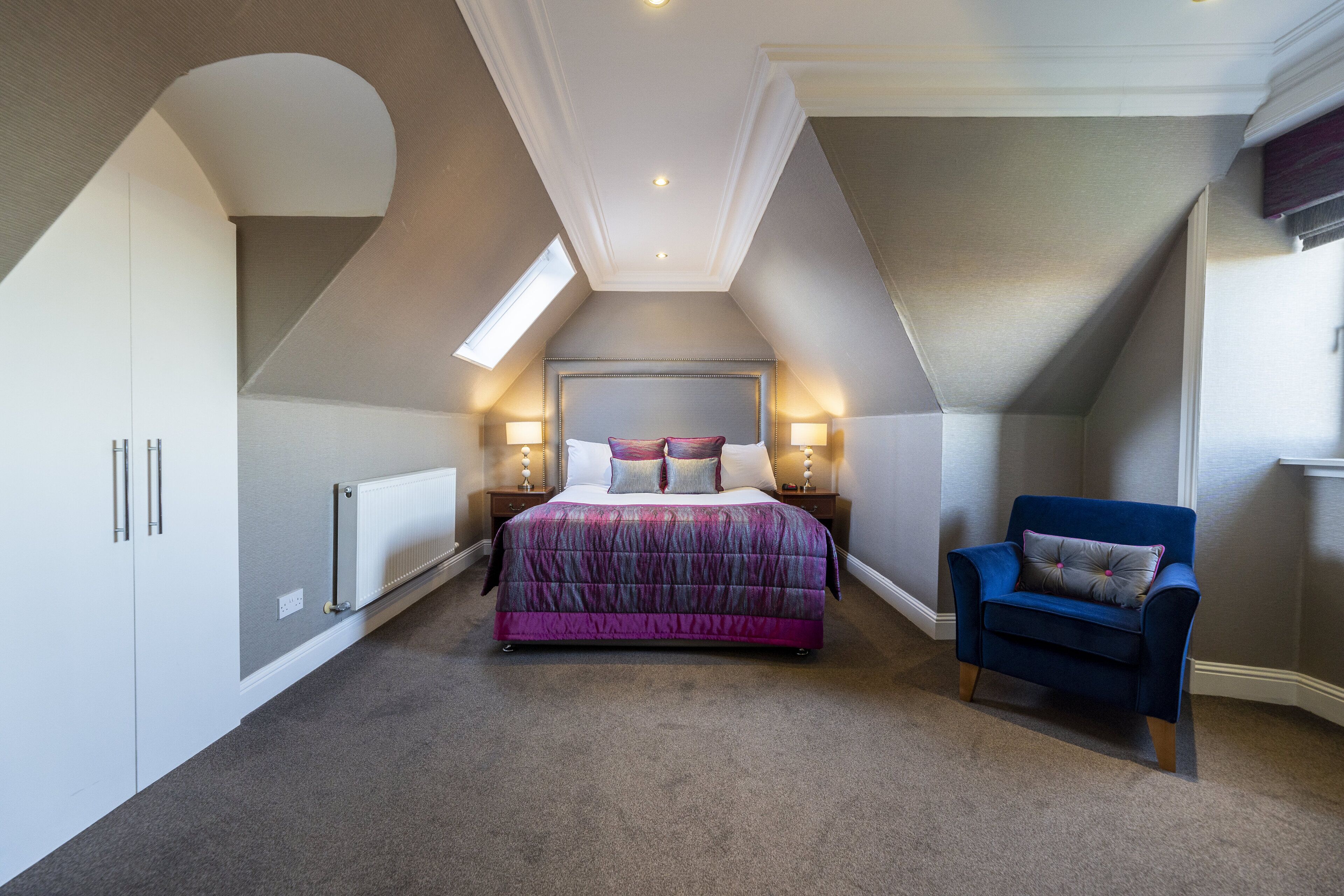 Une chambre avec un plafond en pente, un lit avec une tête de lit, des lampes de chevet, une chaise et un radiateur.