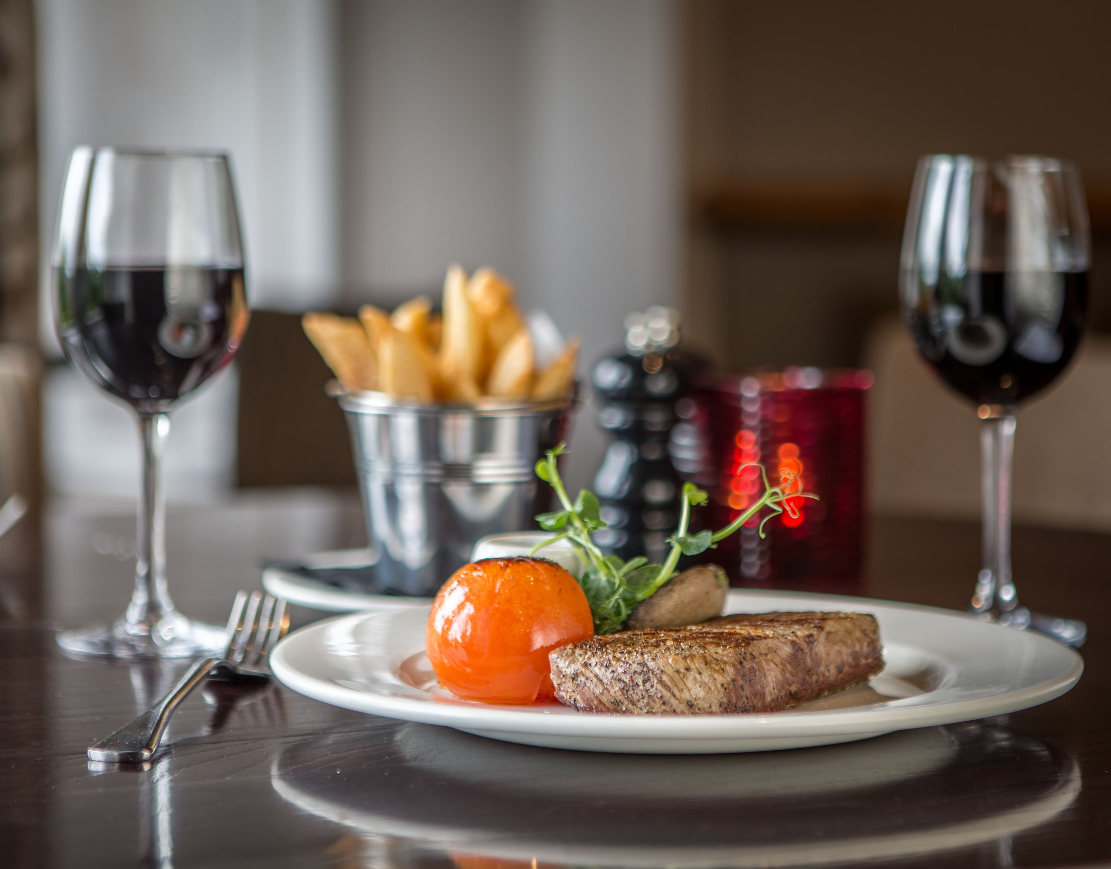 Une assiette contenant un steak, une tomate grillée et une petite salade est posée sur une table. On peut également voir deux verres de vin rouge et un panier en métal rempli de frites.