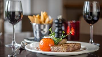 Une assiette contenant un steak, une tomate grillée et une petite salade est posée sur une table. On peut également voir deux verres de vin rouge et un panier en métal rempli de frites.