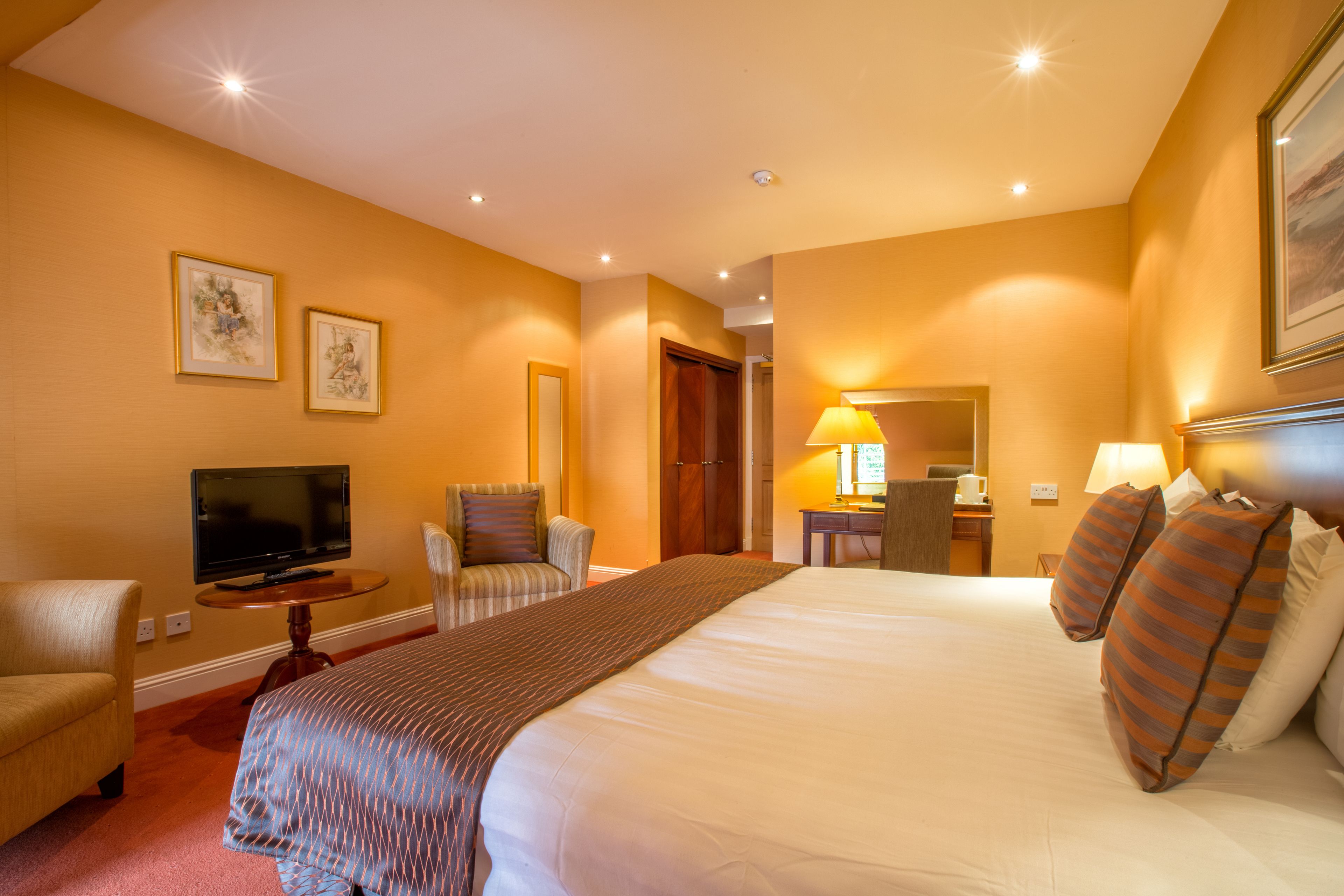 A hotel room with a large bed, a television on a side table, a desk with a lamp, and two framed pictures on the wall.