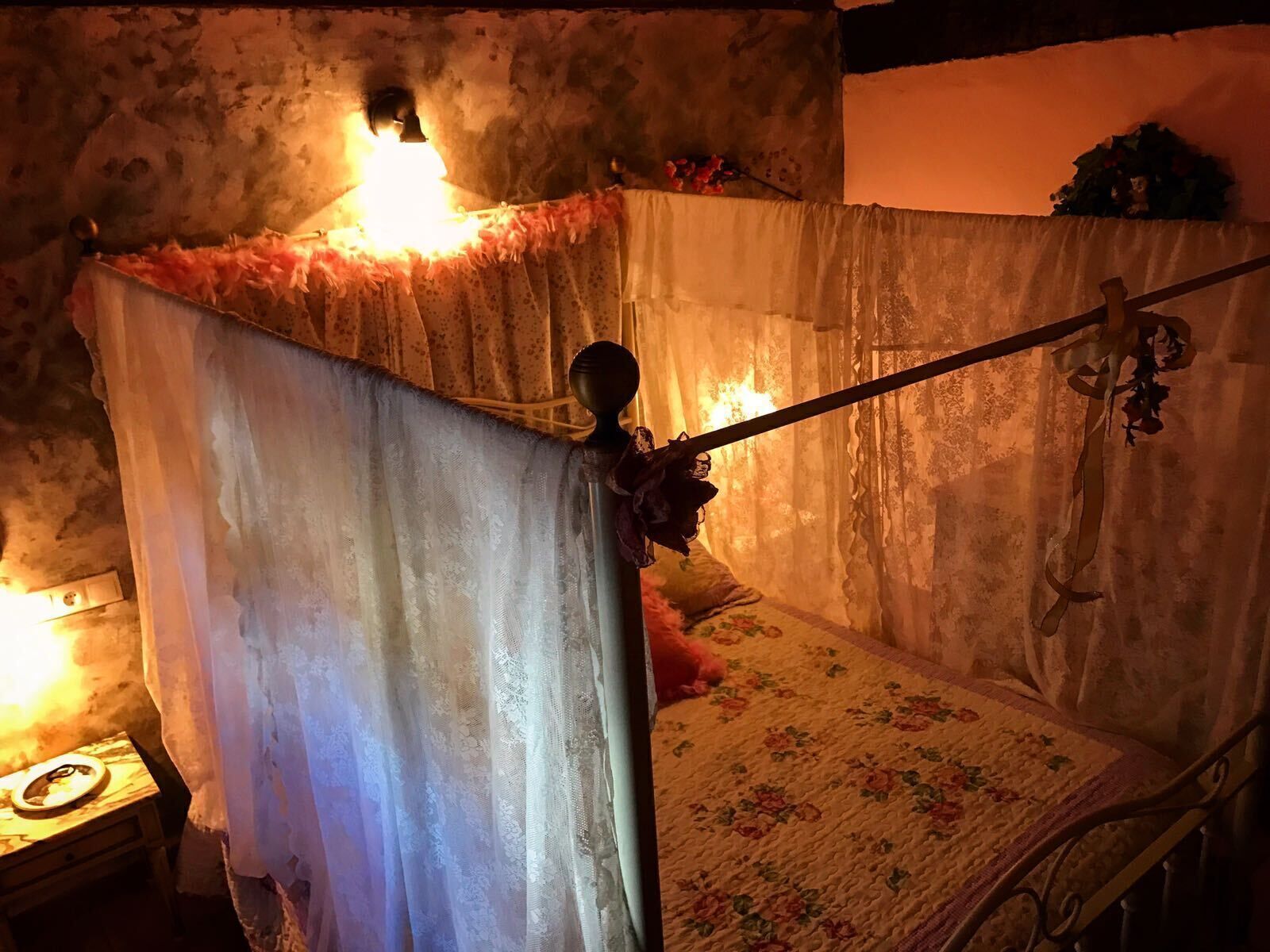 A bed with a canopy, a bedside table with a lamp, and a floral-patterned bedspread.
