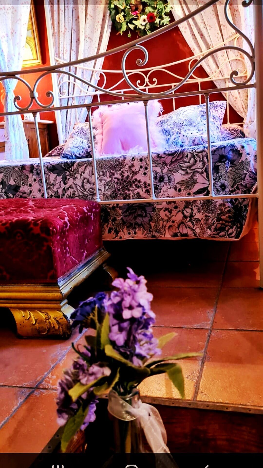 A bedroom with a metal bed frame, floral-patterned bedding, and a vase of flowers on the floor.