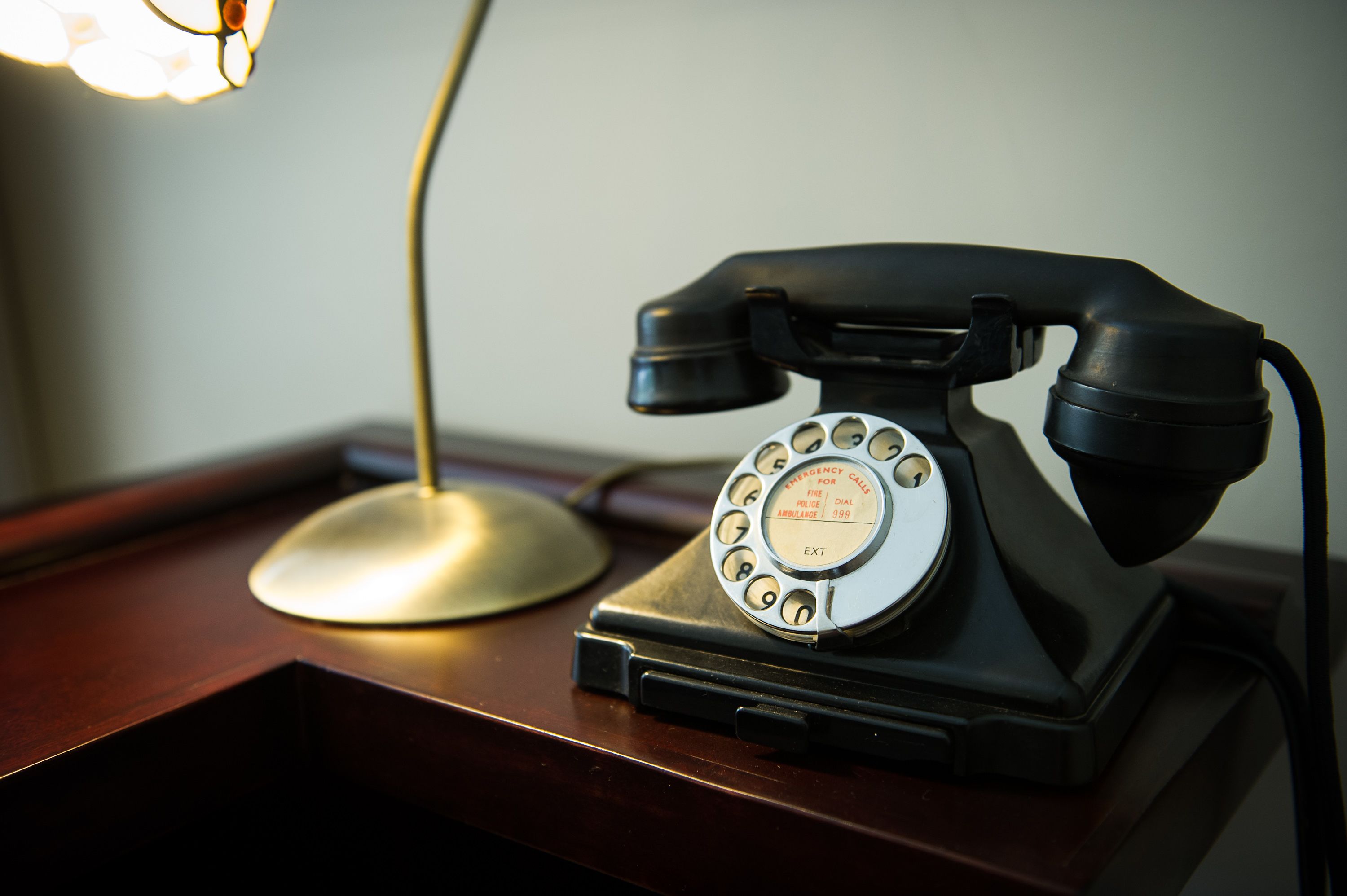 Un teléfono antiguo de disco giratorio sobre un escritorio de madera con una lámpara al fondo.