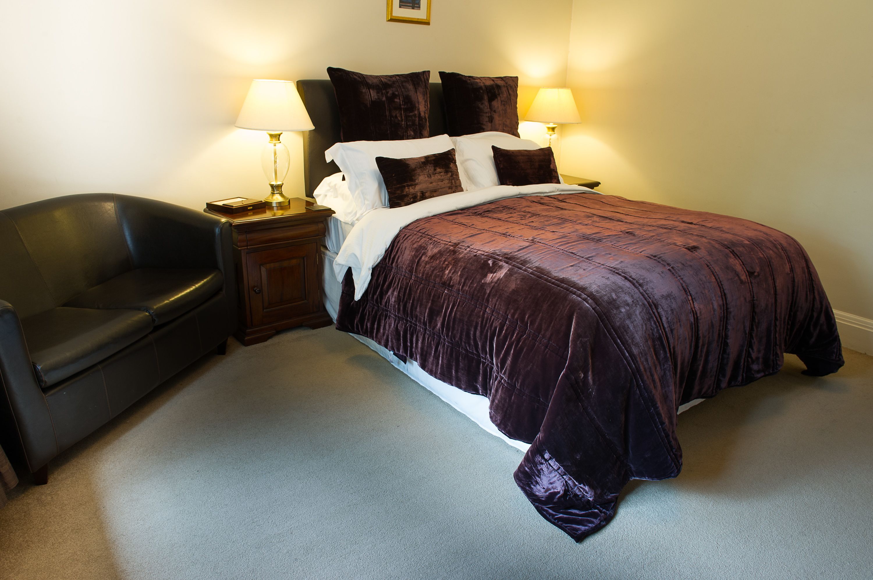 A bedroom with a large bed, two bedside lamps, a dark leather chair, and a wooden nightstand.