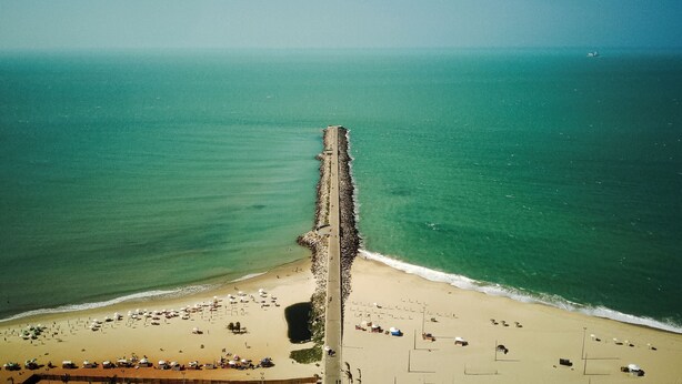 Een luchtbeeld van een strand met een lange pier die zich uitstrekt in de zee, tal van strandparasols en mensen die van het strand genieten.