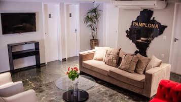 A modern living room with a beige sofa, a glass coffee table with red flowers, a red armchair, a flat-screen TV, and a wall-mounted air conditioner.