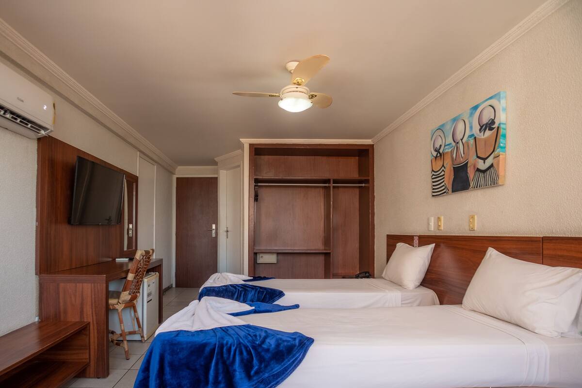 Ein Hotelzimmer mit zwei Betten, einem Schreibtisch, einem Stuhl, einem Fernseher, einem Deckenventilator und einem Kleiderschrank.