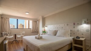 Een hotelkamer met een bed, een bureau, twee stoelen, een nachtkastje met een lamp, en uitzicht op gebouwen door het raam.