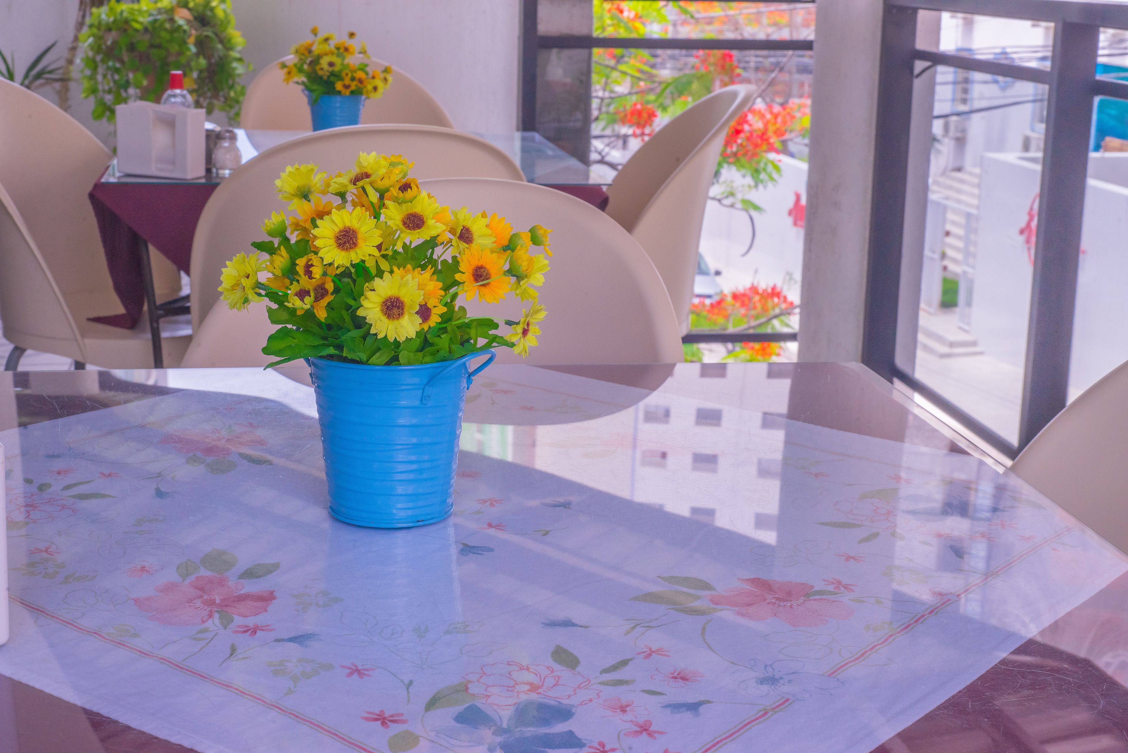 A table with a floral tablecloth, a blue vase with yellow flowers, and beige chairs.