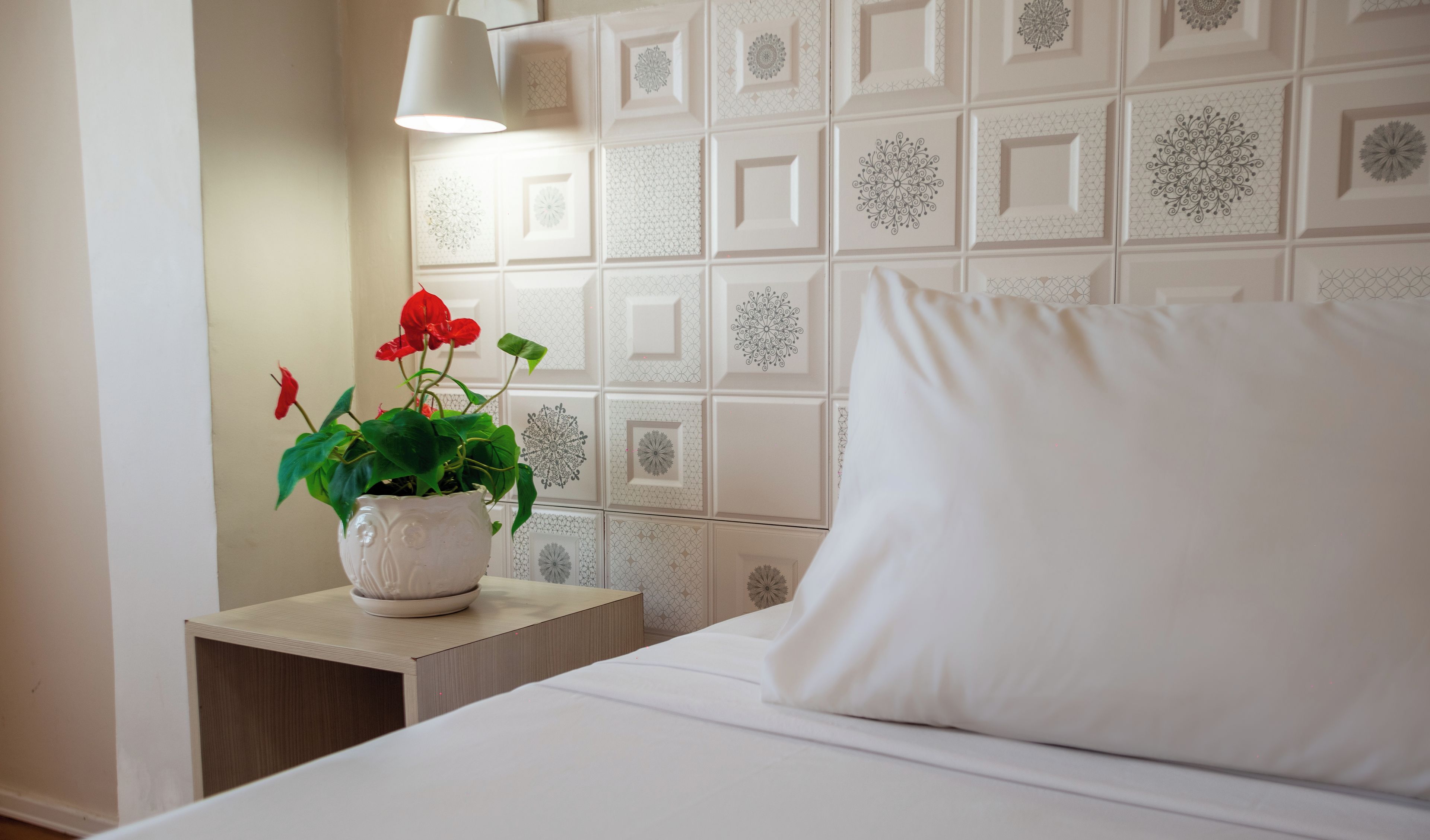 Uma cama bem arrumada com um travesseiro branco e uma pequena mesa de cabeceira branca ao lado. Um vaso branco com uma flor vermelha está na mesa de cabeceira.