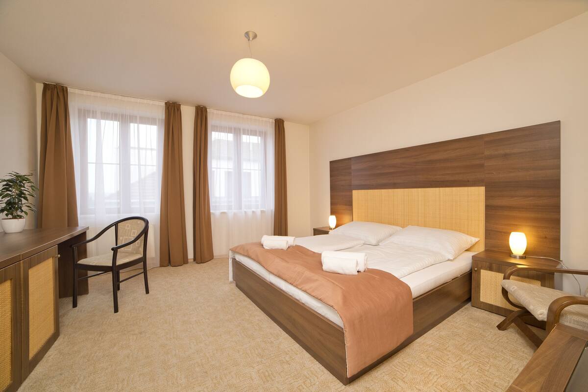 A hotel room with a bed, bedside tables, a desk, a chair, and a potted plant.