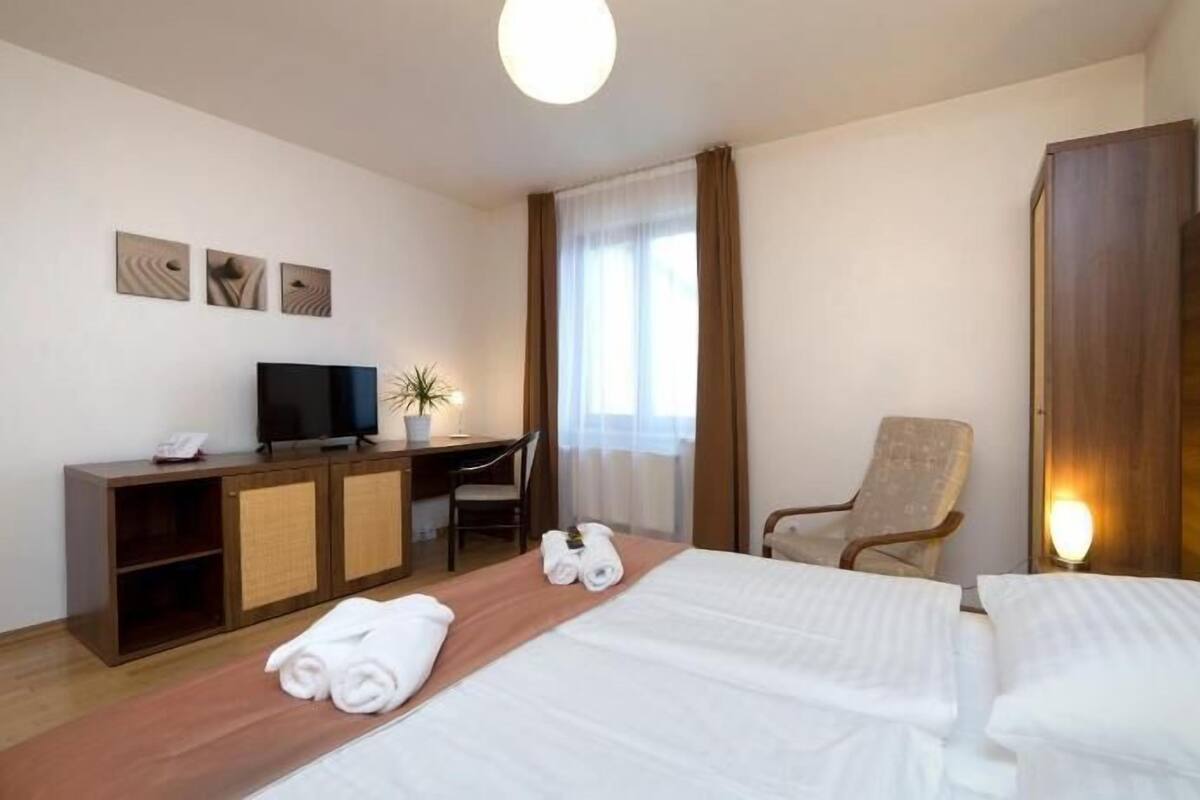 A hotel room with a bed, a desk with a TV, a chair, and a window with curtains.