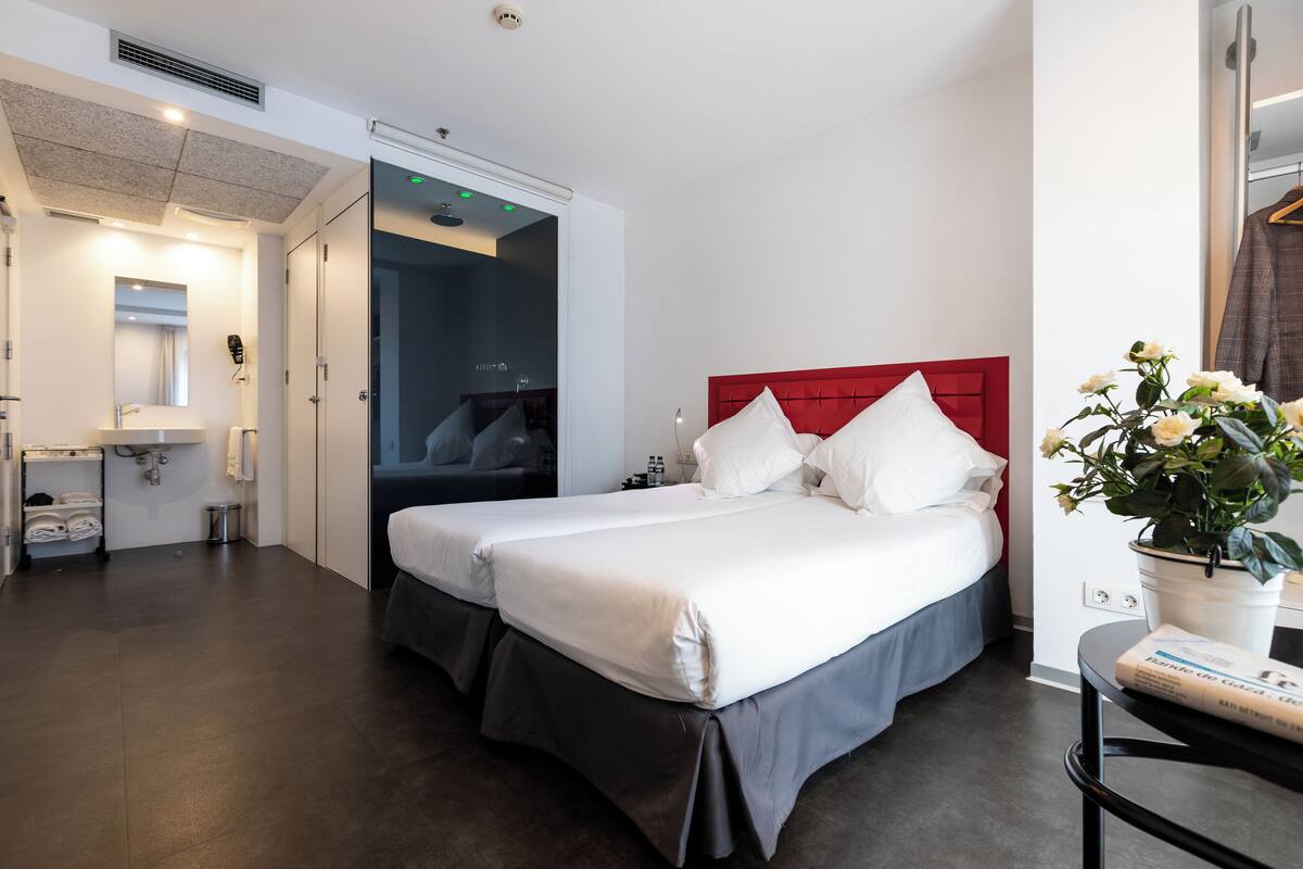 A modern hotel room with a large bed, a red headboard, a small bedside table, and a potted plant.