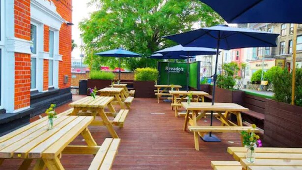 An outdoor patio with wooden tables and benches, covered by blue umbrellas, and a sign for 'Ravado's' in the background. - Revado Hotel, Norwich City Centre (Norwich)