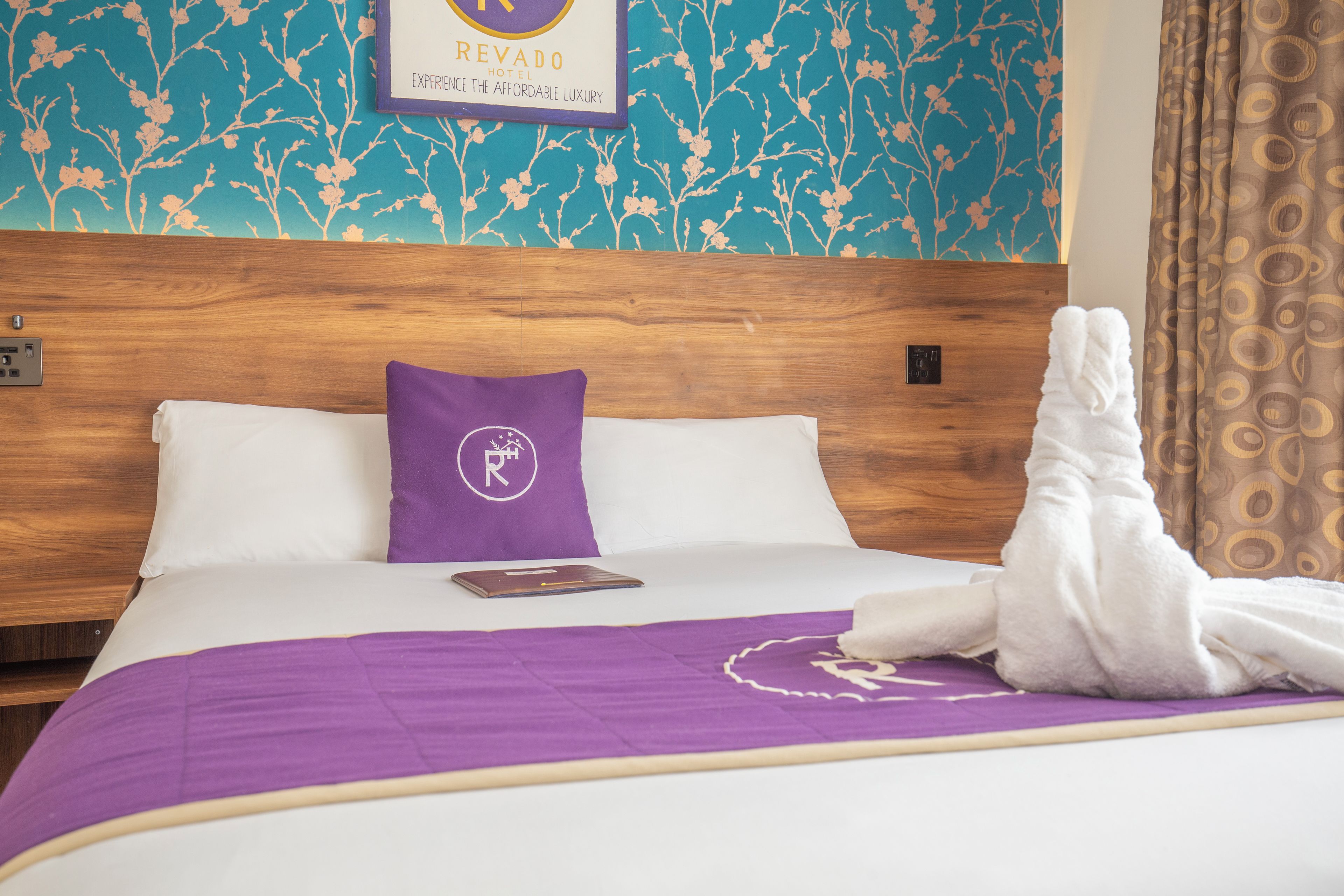 Een hotelkamer met een houten hoofdeinde, een paars kussen met een logo, een witte badjas en een paars sprei.