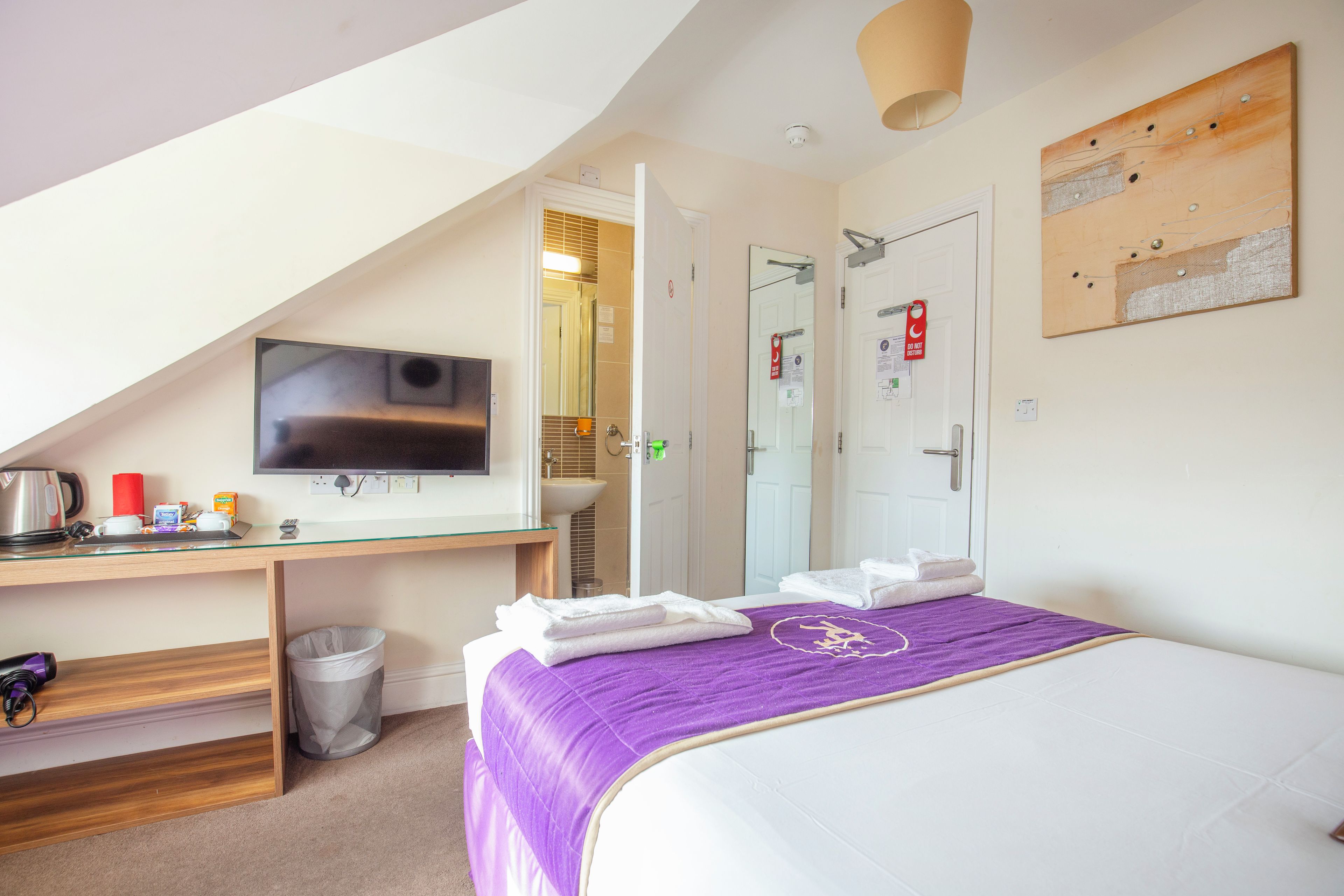 A hotel room with a bed, a television, a desk with a kettle, and a trash bin.