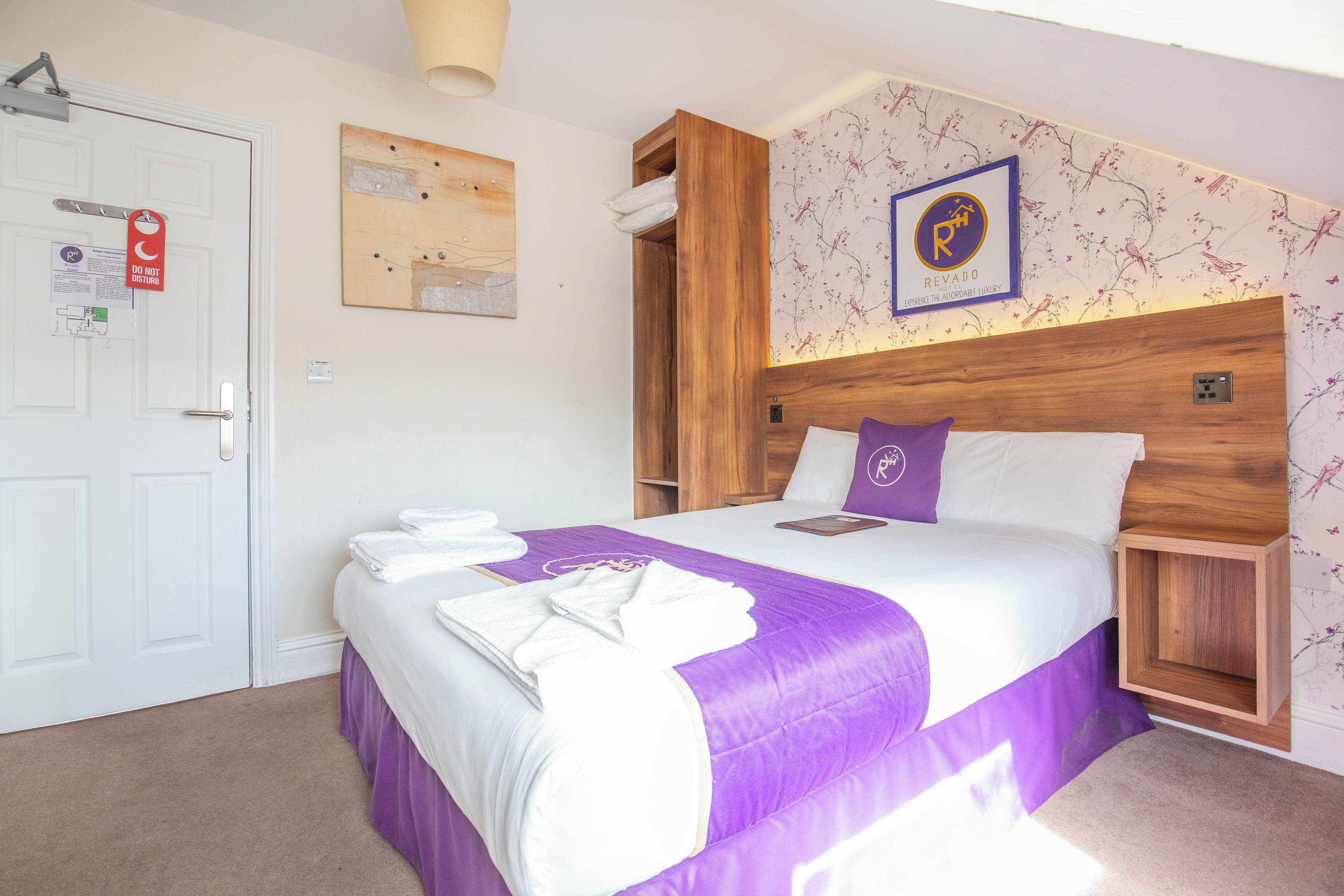 Habitación de hotel con una cama, cabecera de madera, un cuadro colgado en la pared y una puerta con el cartel de 'No molestar'.
