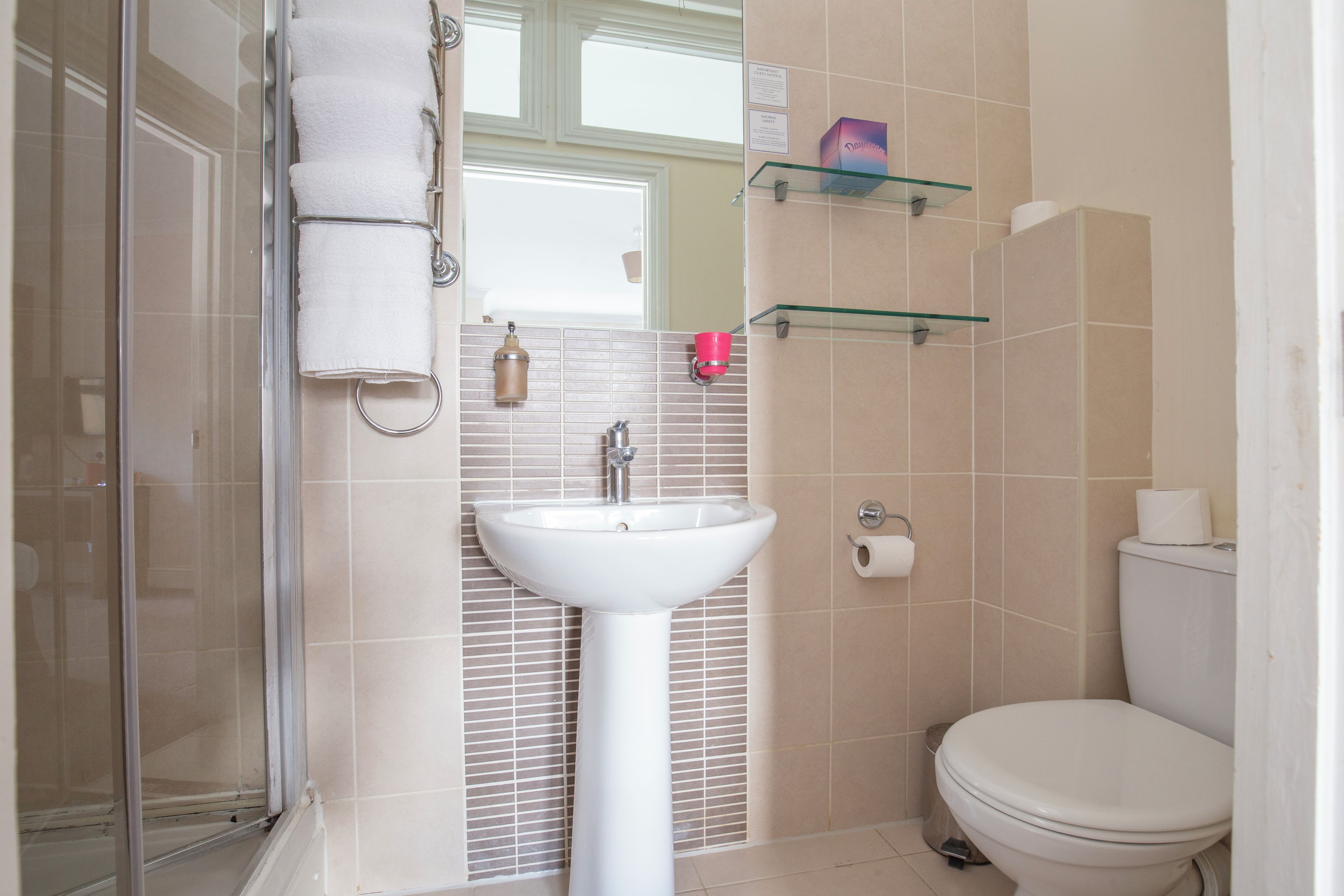 Une salle de bain avec une douche à parois de verre, un lavabo blanc, des toilettes et une étagère en verre où sont rangés les articles de toilette.
