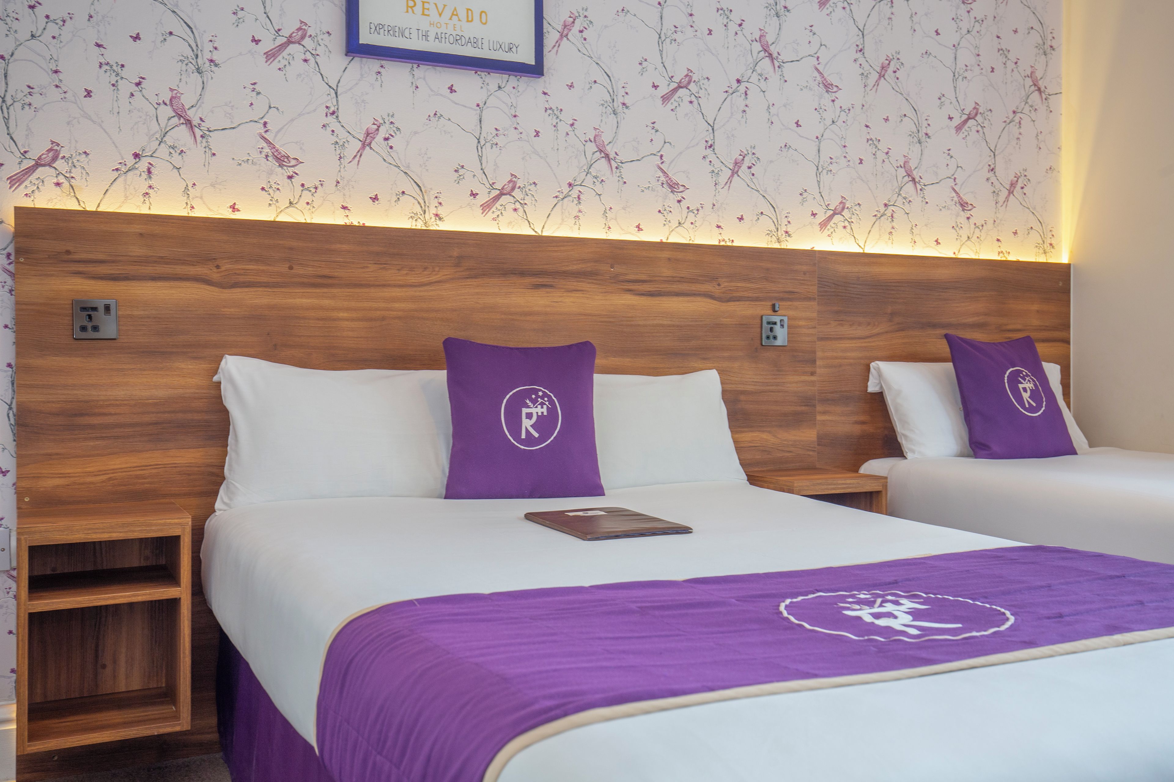 Habitación de hotel con dos camas, cada una con cabecera de madera, ropa de cama blanca y almohadas moradas con un logotipo. Se observa una mesita de noche con una lámpara y un enchufe en la pared.