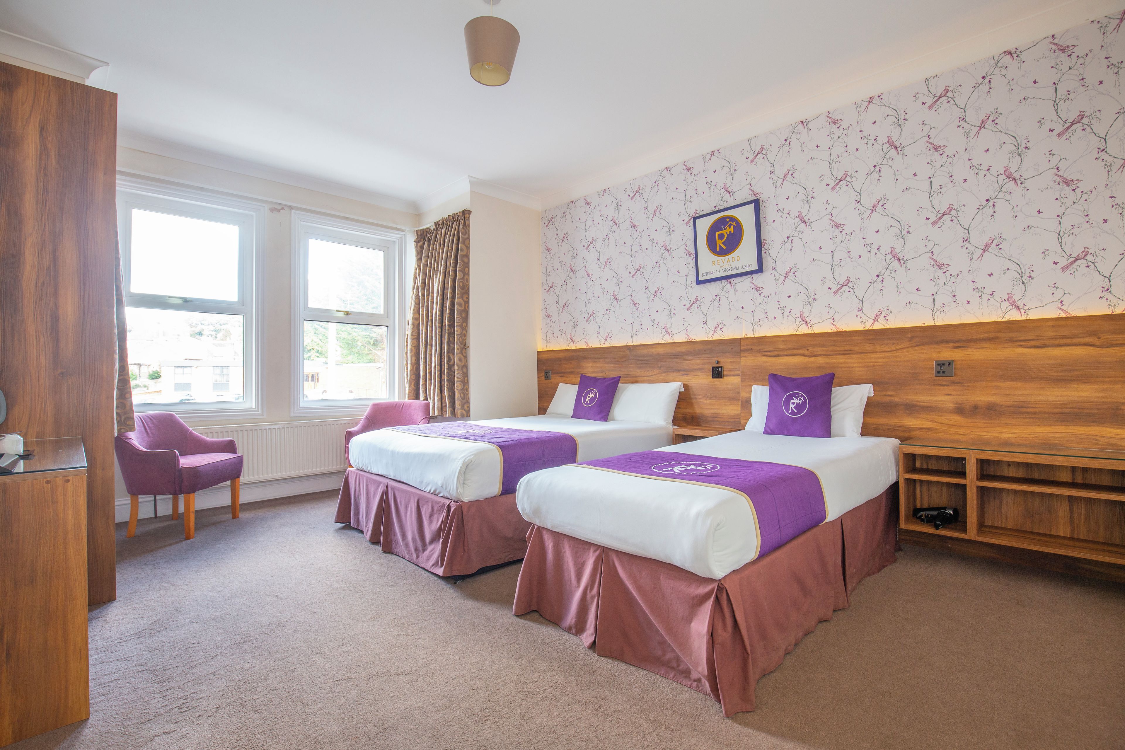 A hotel room with two beds, a wooden headboard, a window with curtains, a chair, and a wall with a bird pattern.