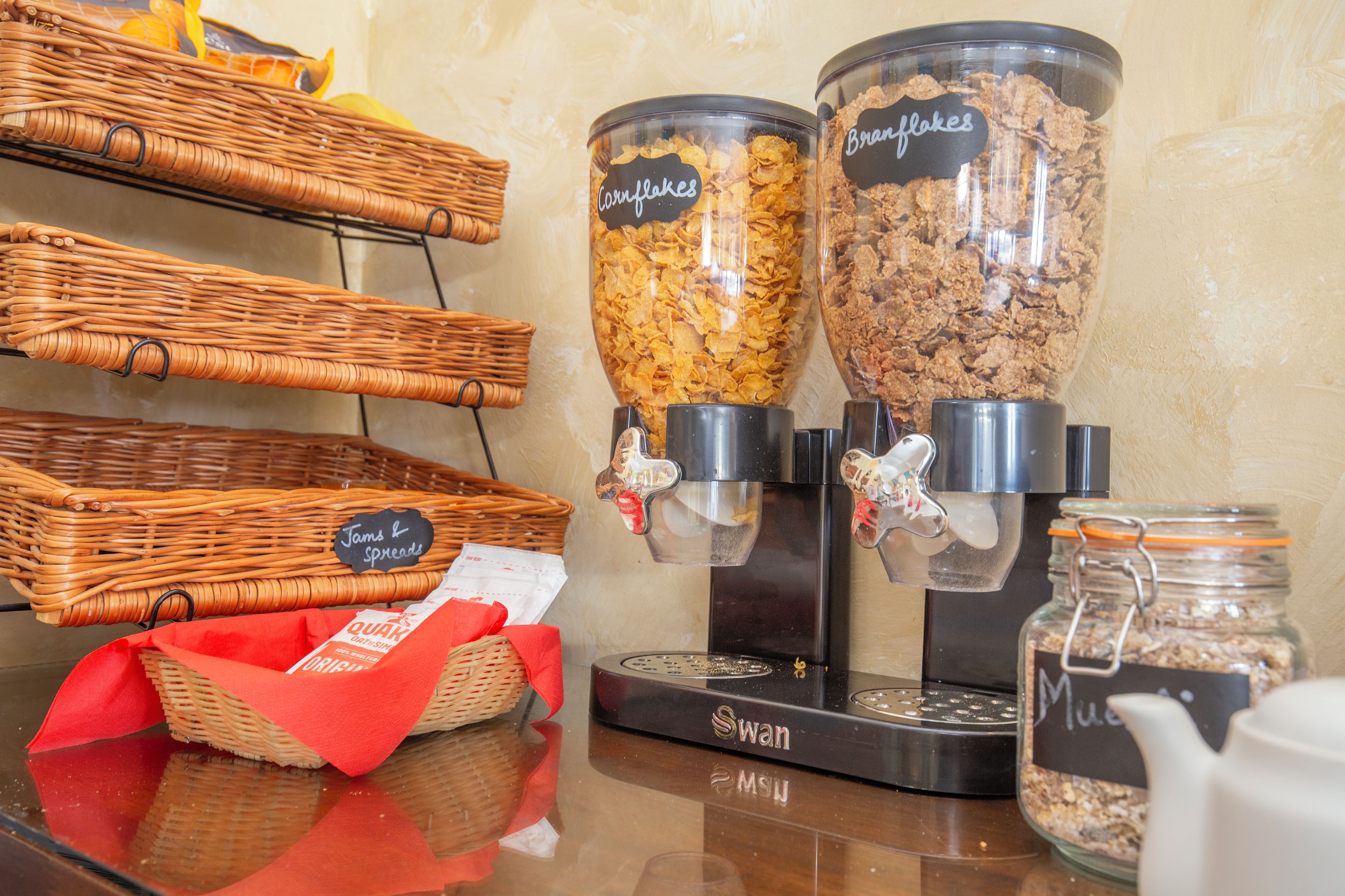 A self-serve coffee station with labeled dispensers for cornflakes and bran flakes, a wicker basket, and a jar of muesli.