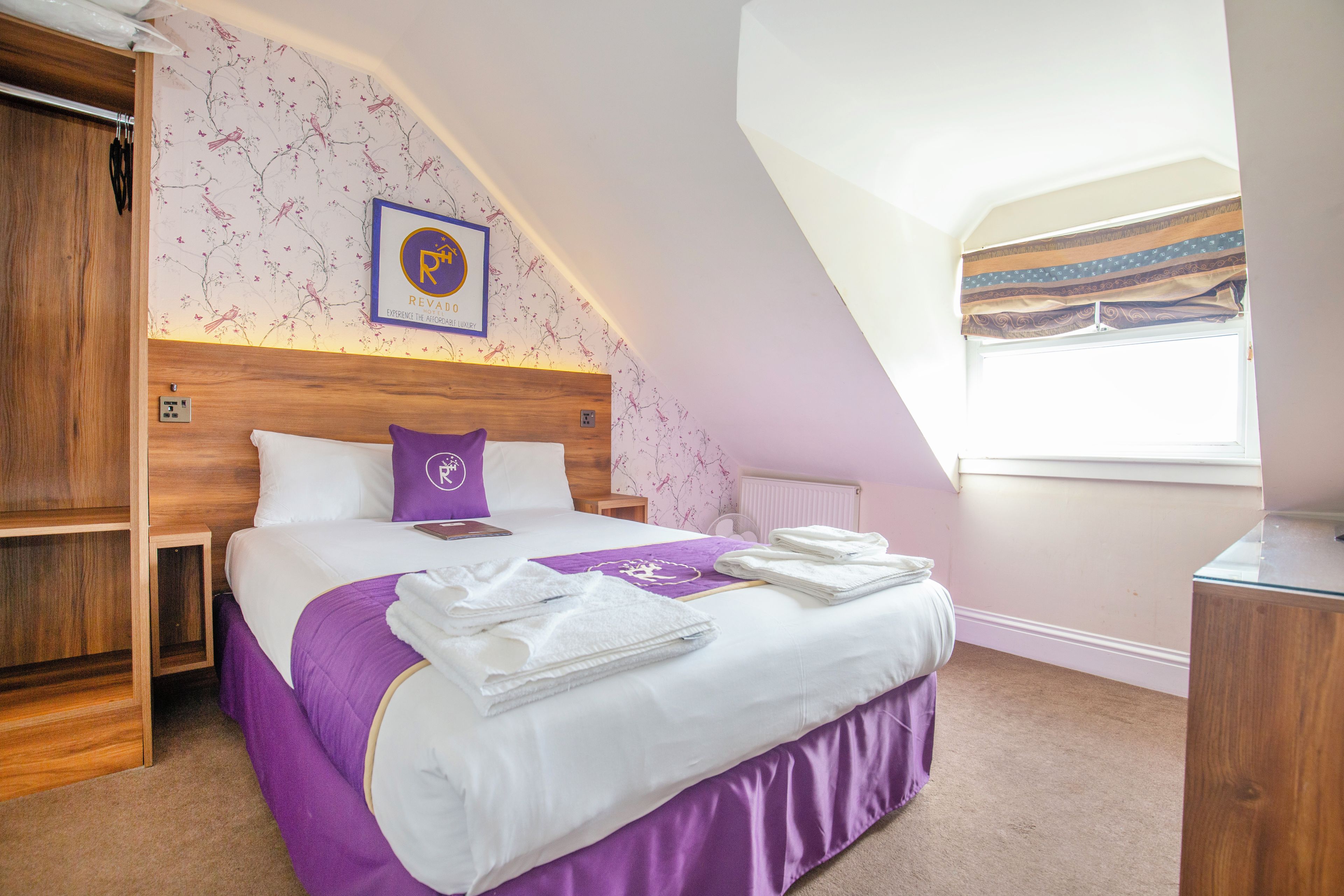 Un dormitorio con cabecera de madera, cama con ropa de cama blanca y morada, ventana con cortinas y pared con papel tapiz floral.