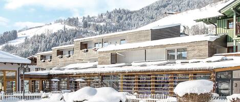 A snow-covered building complex with multiple structures, a white picket fence, and a mountainous backdrop.