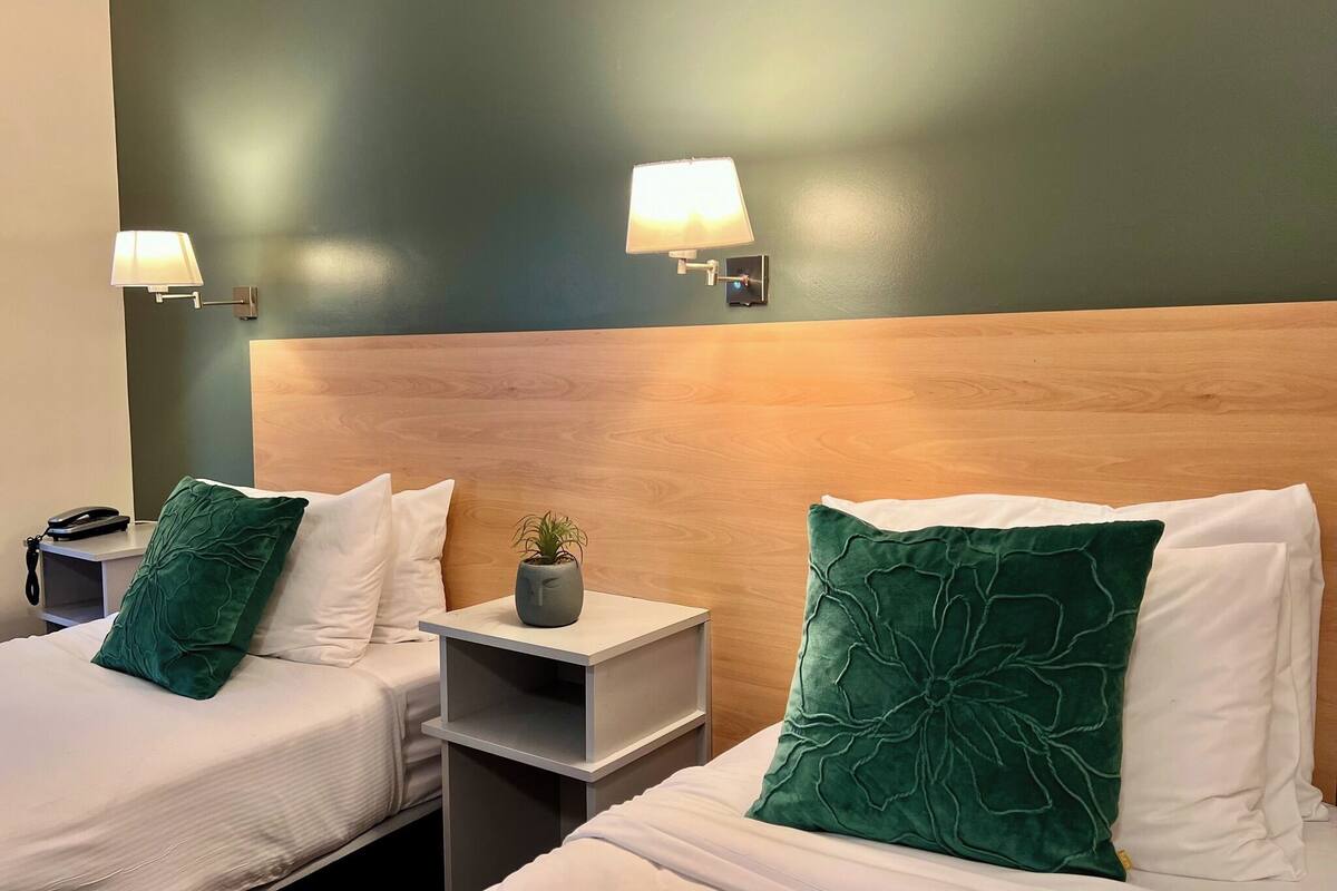 Ett hotellrum med två sängar, var och en med en grön kudde, ett sängbord med en lampa och en liten växt på bordet.
