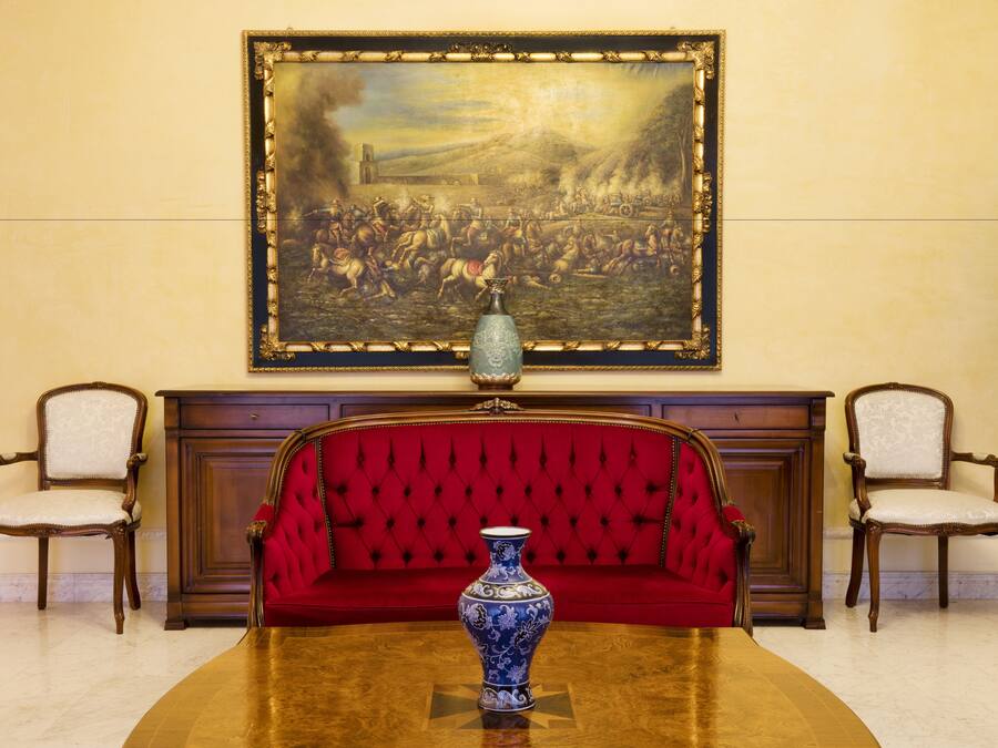A formal room with a red upholstered sofa, a wooden table, and a large framed painting above the fireplace.